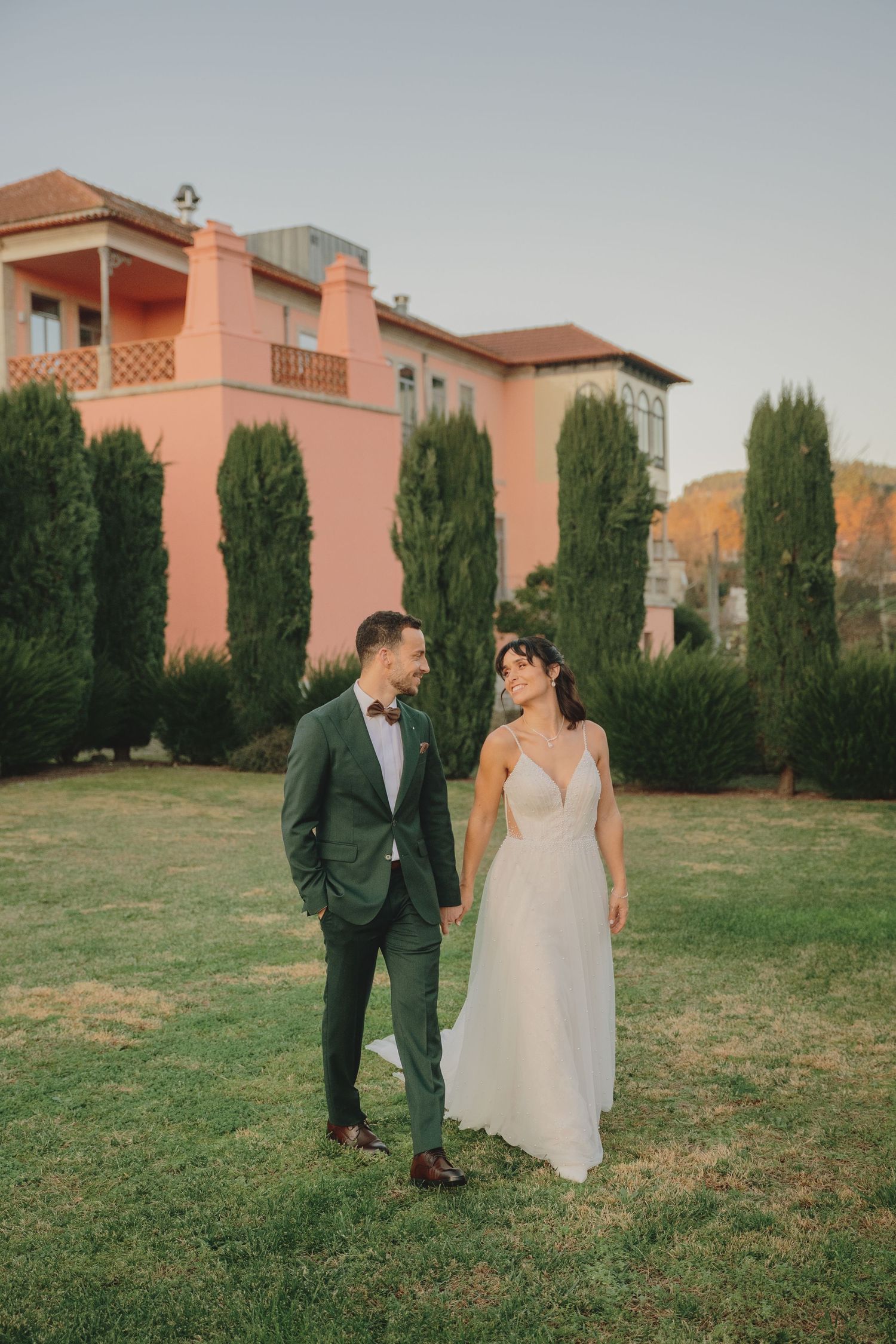 Wedding couple walking on grass near pink Mediterranean-style buildings.