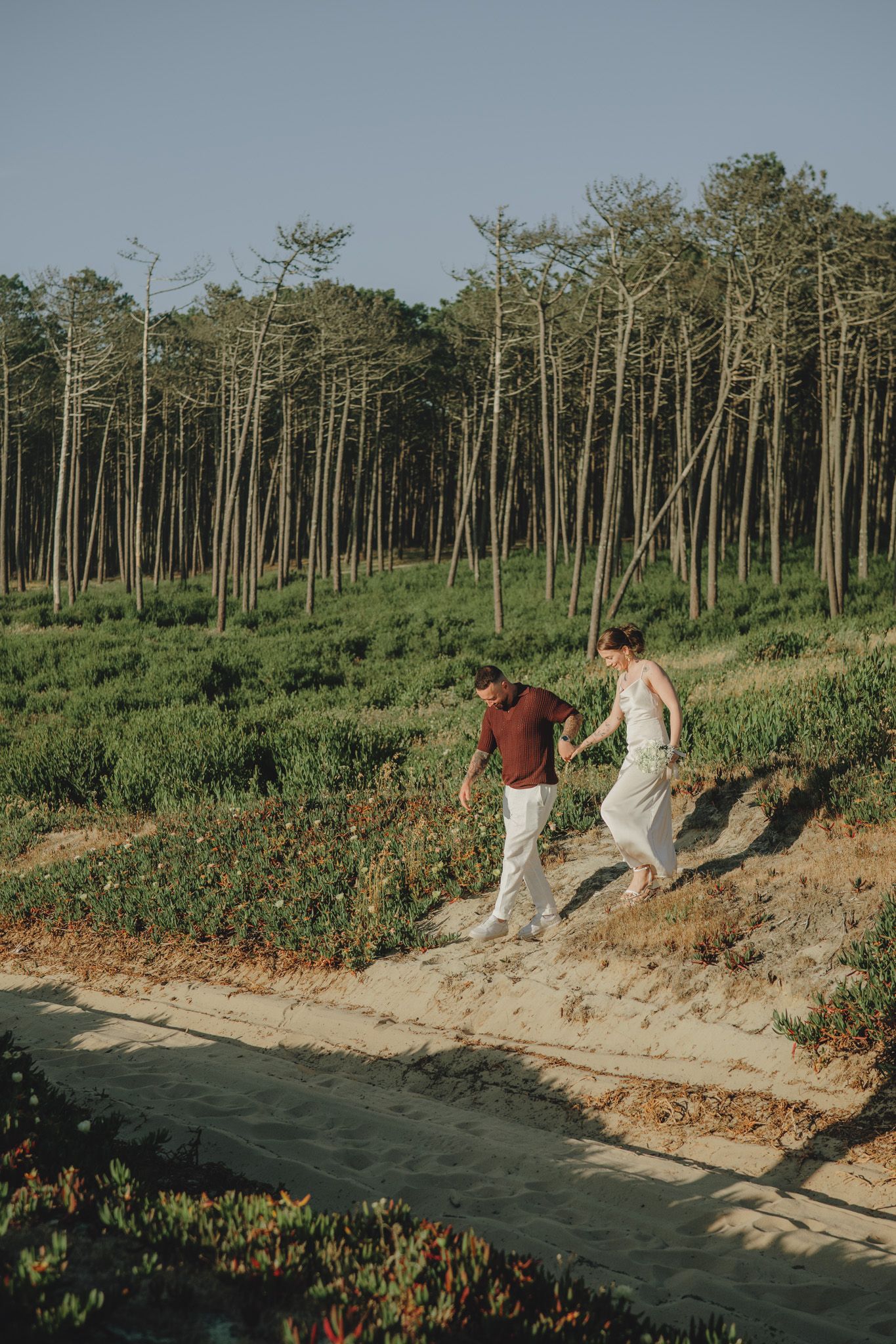 Two people in white outfits walking along a scenic coastal path with tall trees in the background.