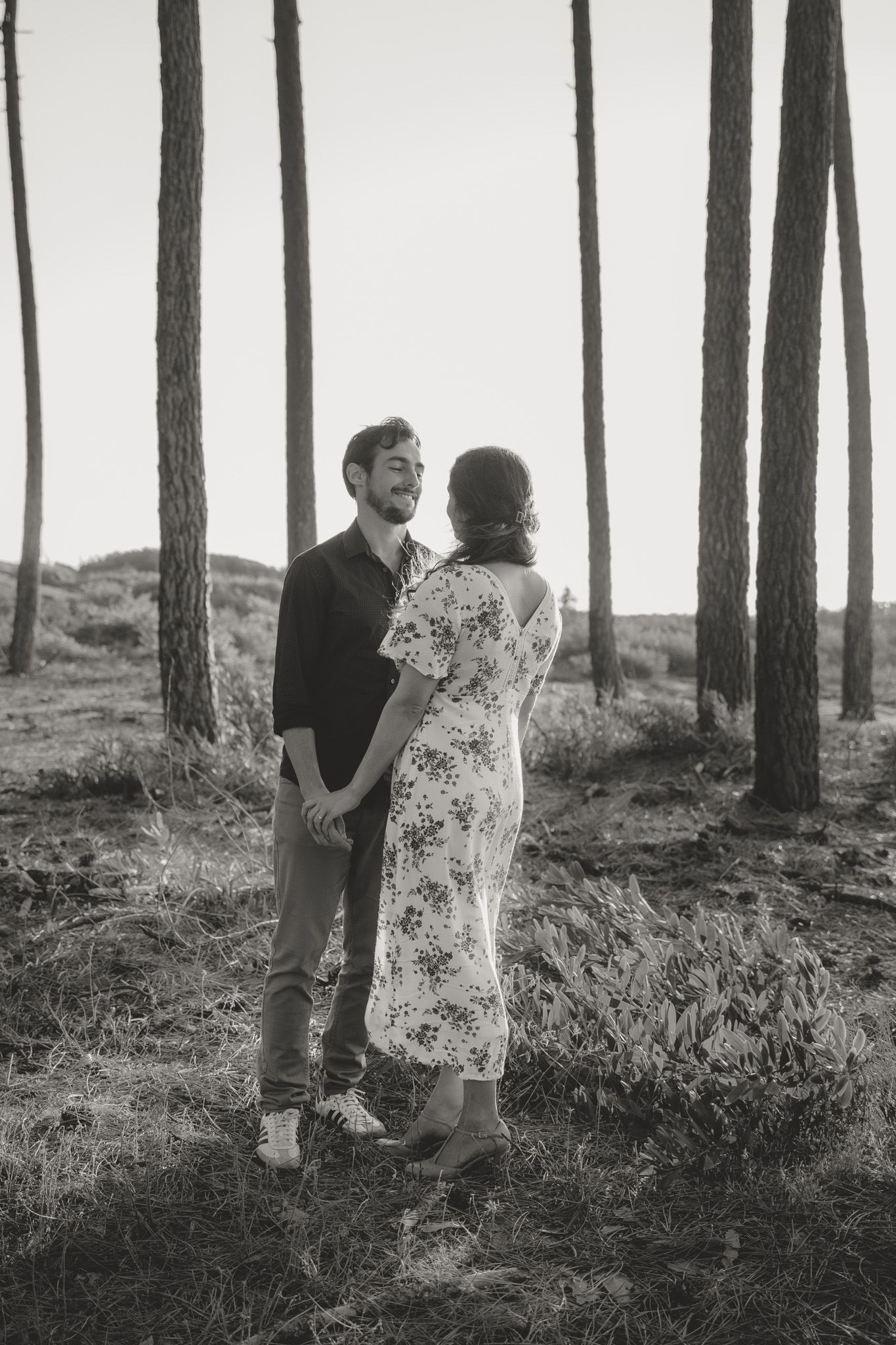 A couple shares a romantic moment among tall pine trees in black and white.