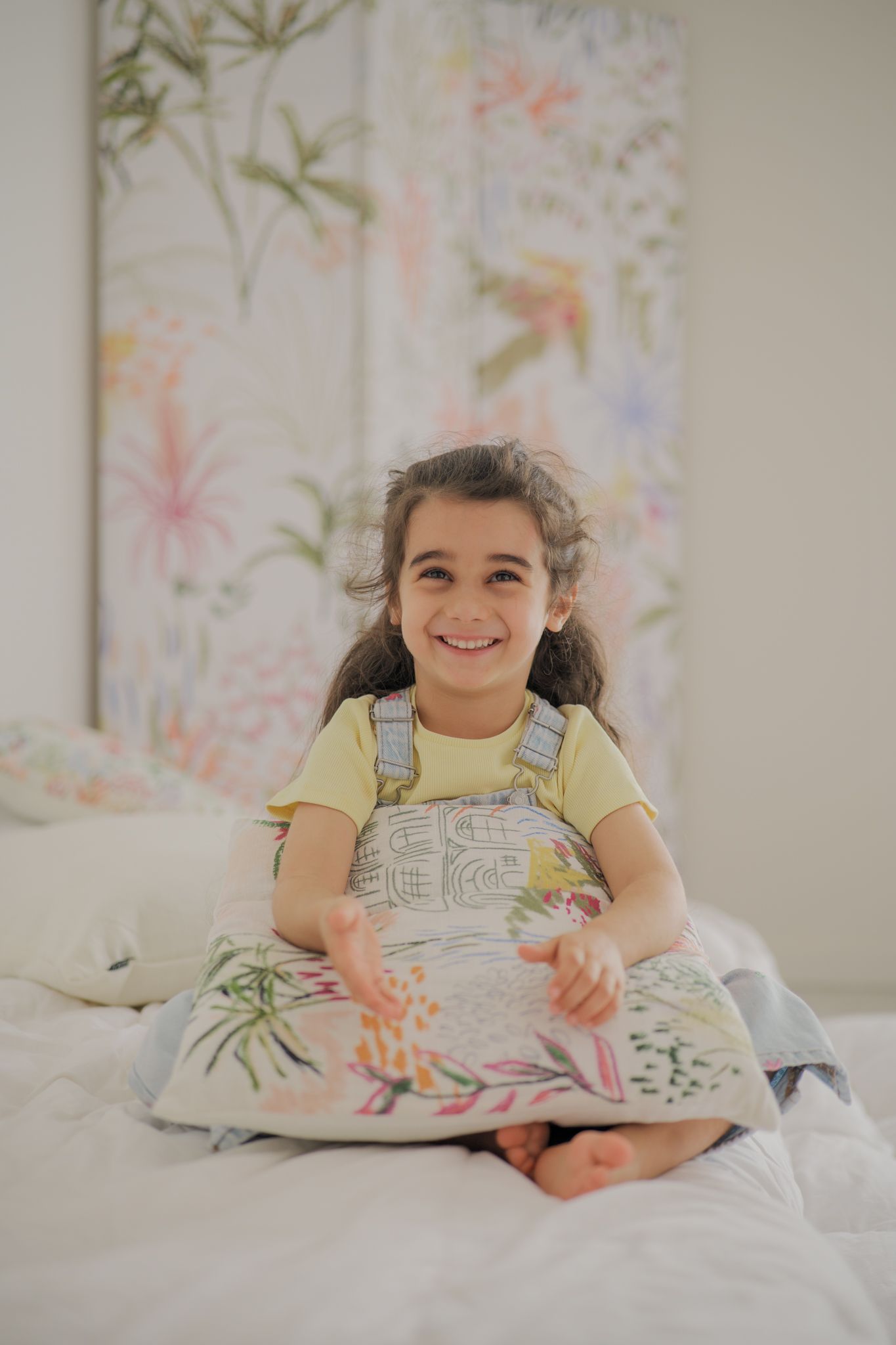 Smiling young girl sitting on a white bed holding a floral pillow, wearing a yellow shirt and denim overalls.