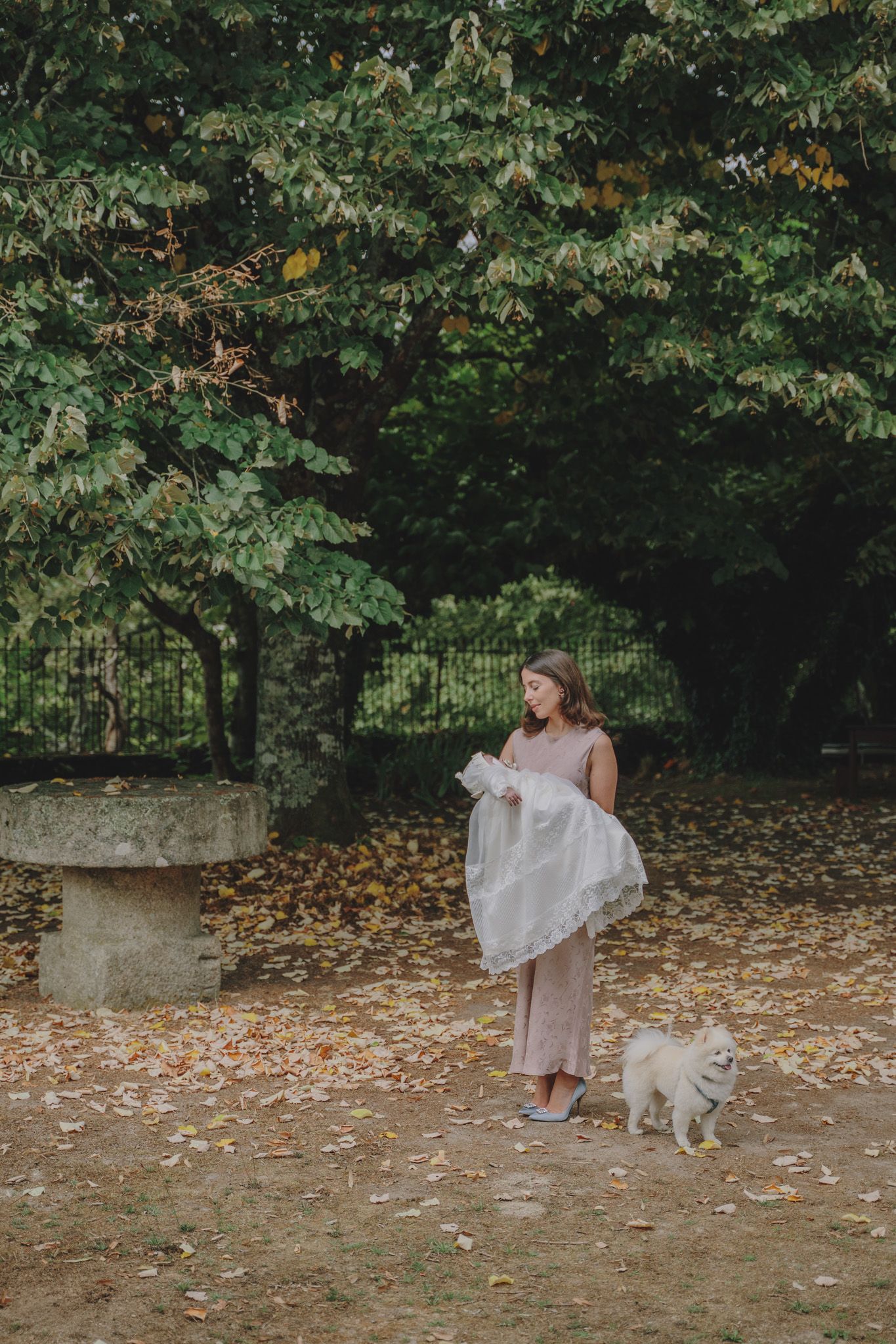Figure in a light dress walks with a small white dog through an autumn garden with fallen leaves.