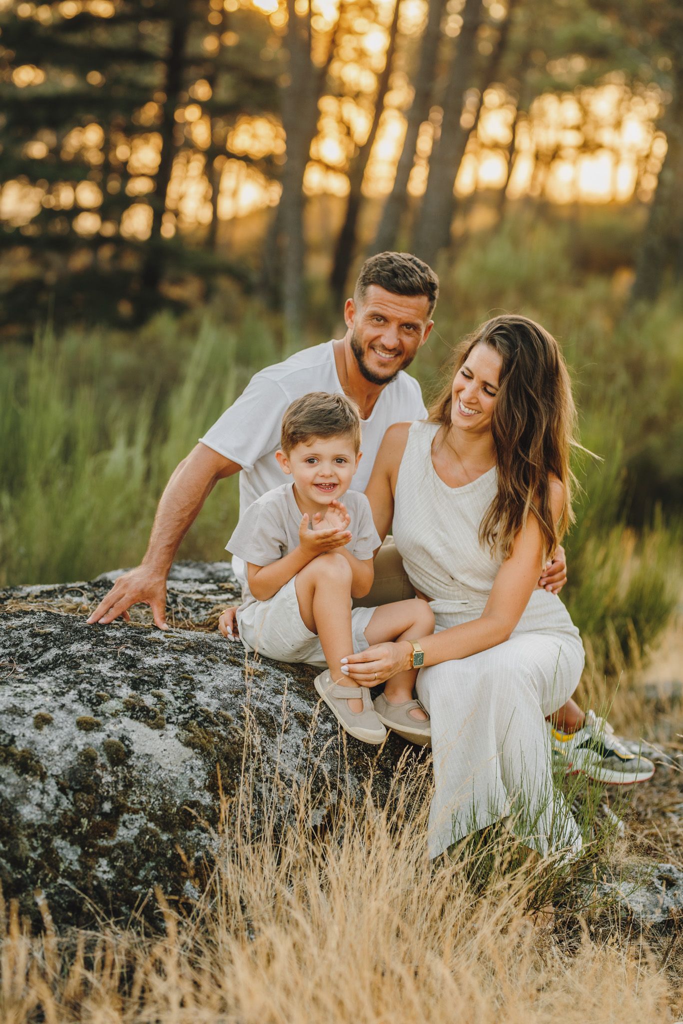 Family enjoying sunset together on rocky terrain surrounded by tall grass and pine trees.