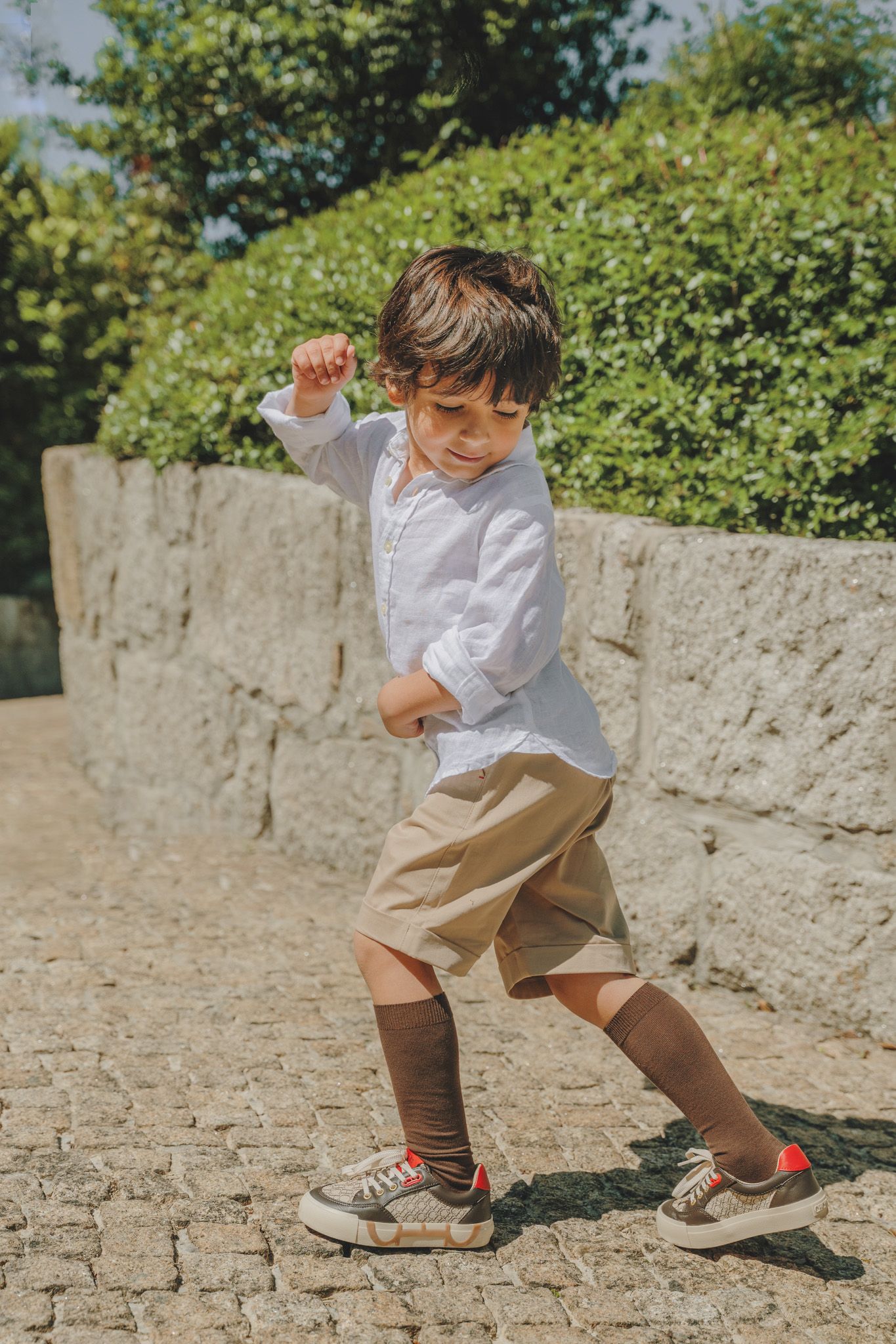 A child in white shirt and khaki shorts jumping playfully on cobblestone pavement.