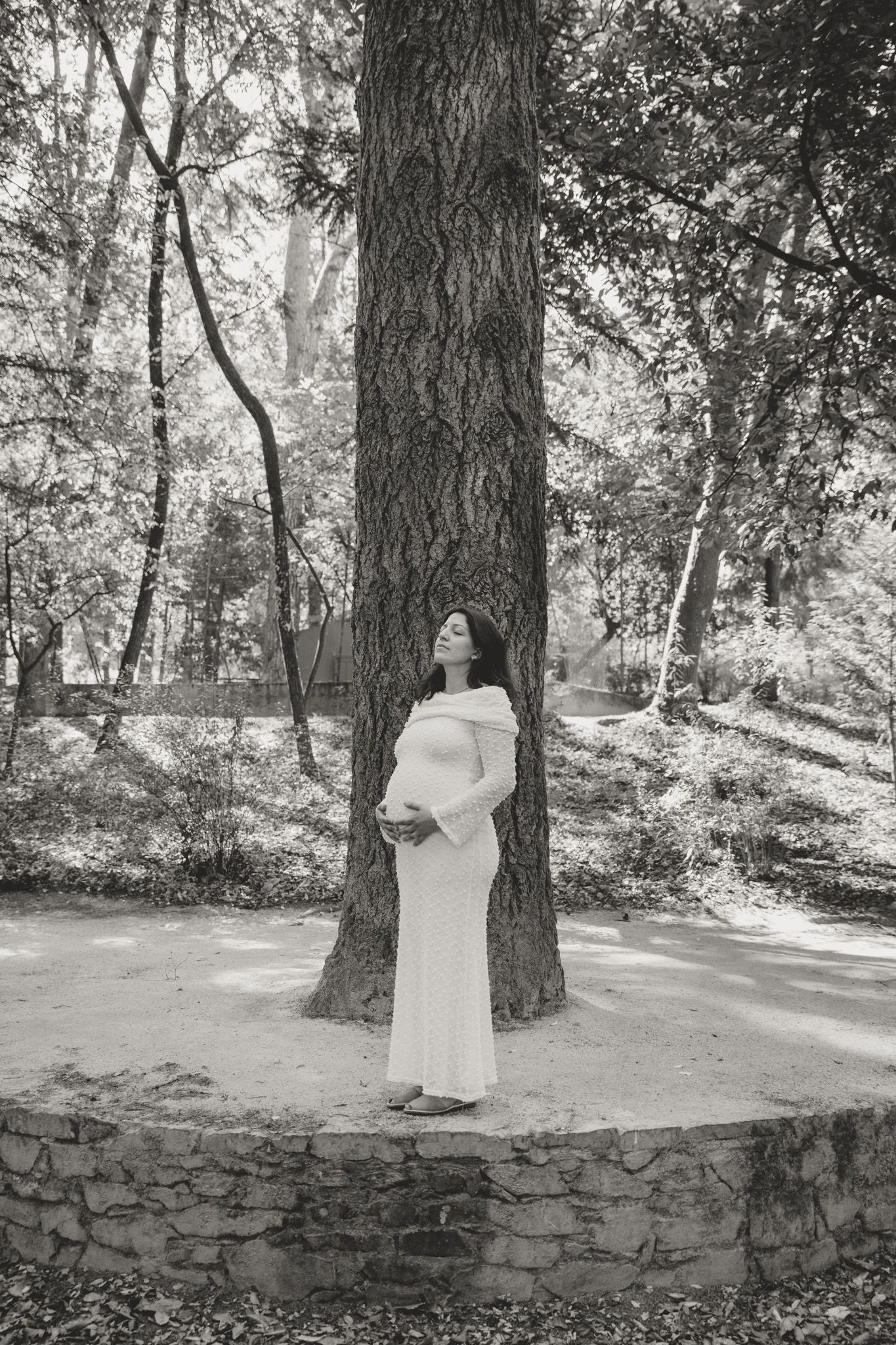 Person in white dress poses against stone wall in wooded setting.