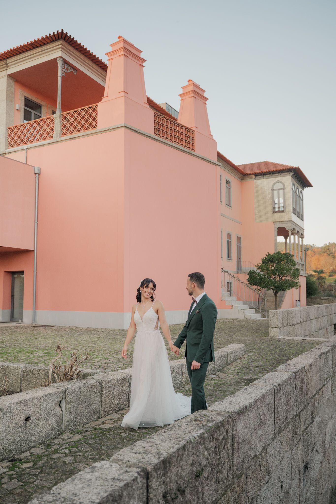Wedding couple walks along a pink Mediterranean villa wall during golden hour photo session.