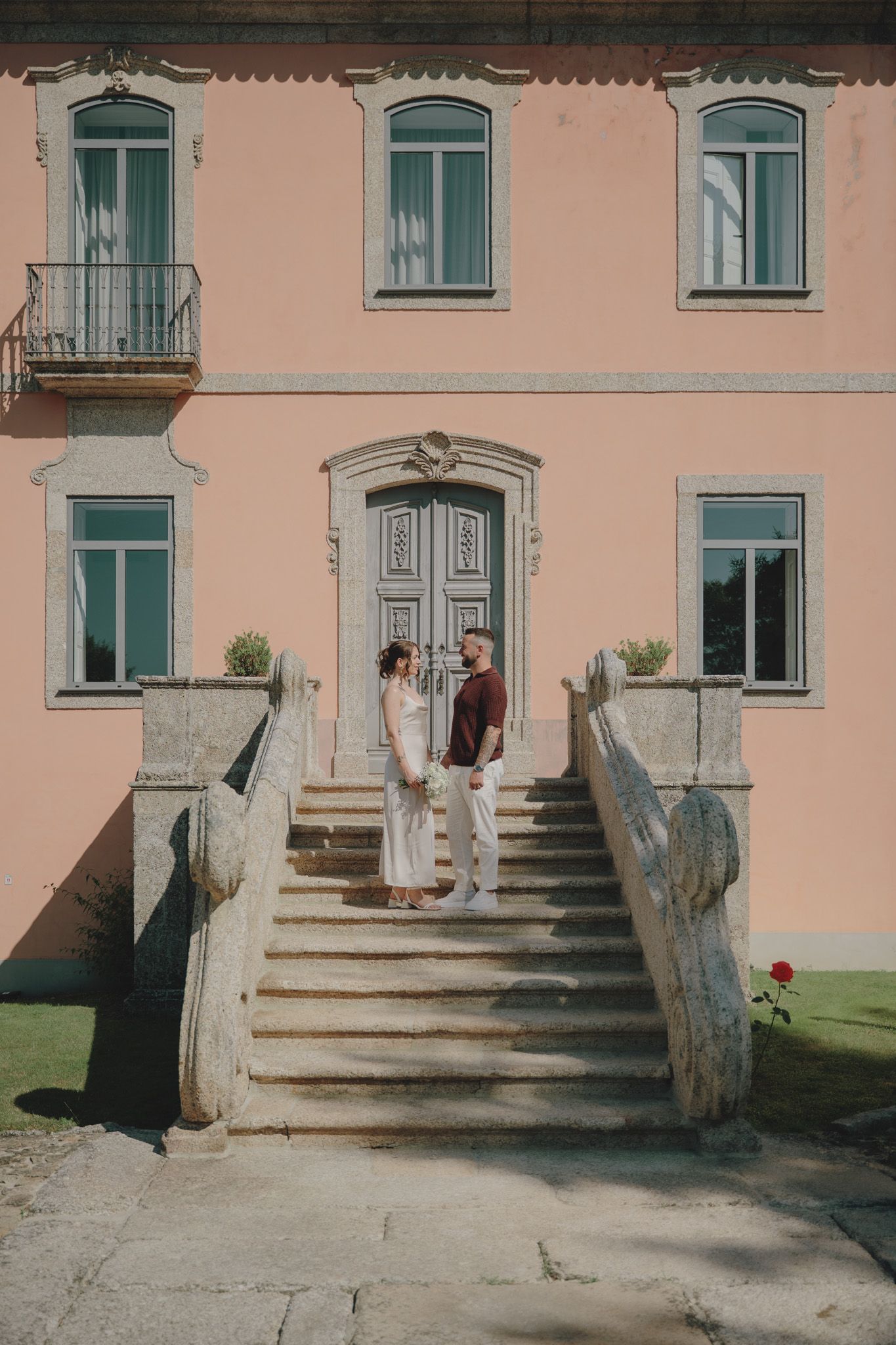 A couple stands on stone steps in front of a pink building with arched windows.