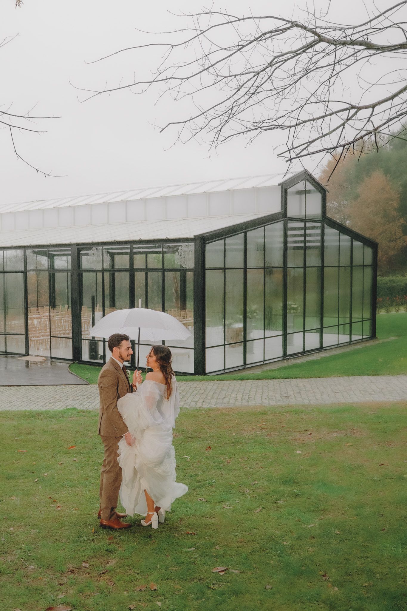 Modern glass greenhouse venue with couple dancing on lawn during autumn wedding.