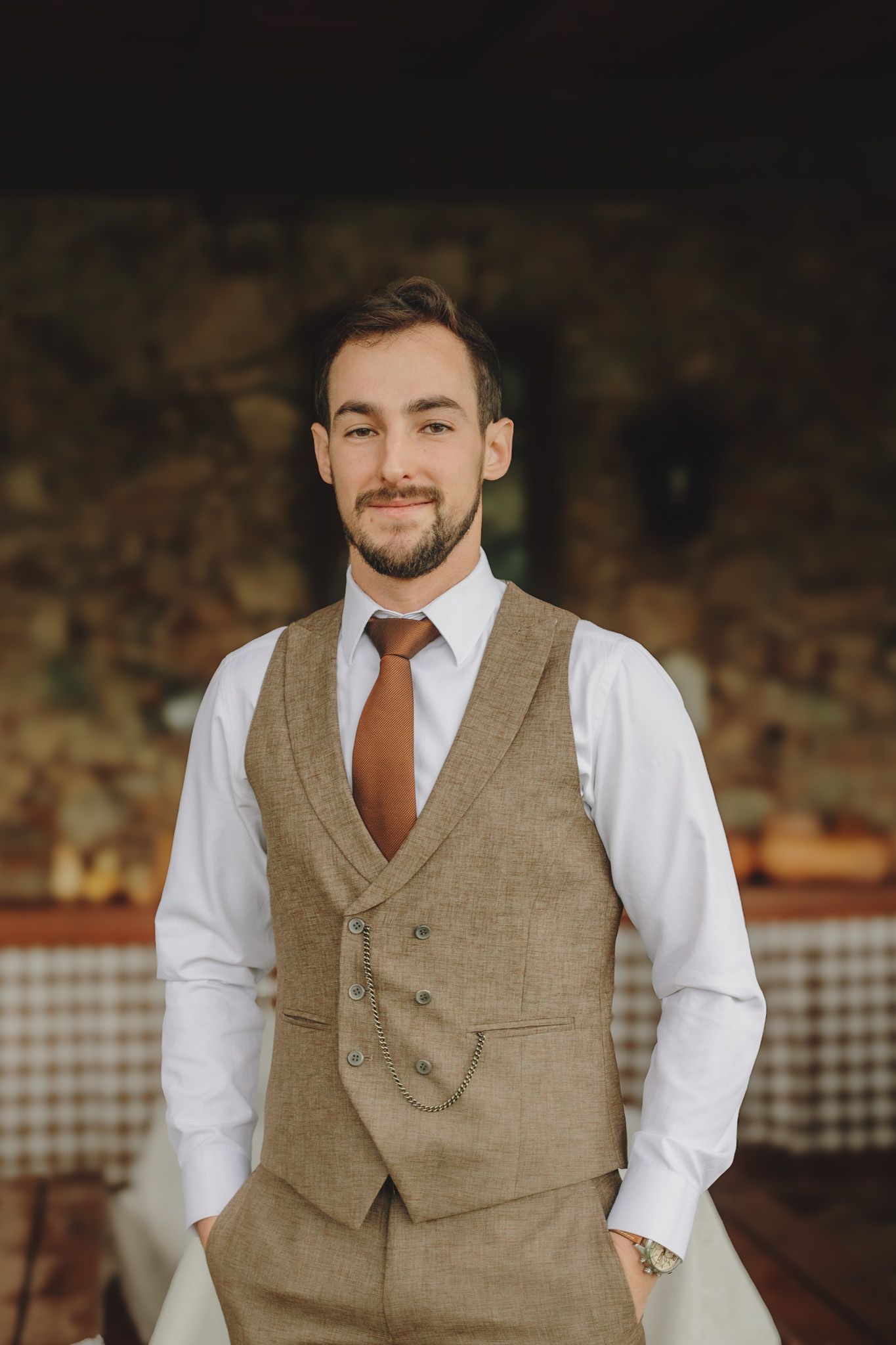 Groom wearing a brown tweed vest and white dress shirt in a rustic setting.