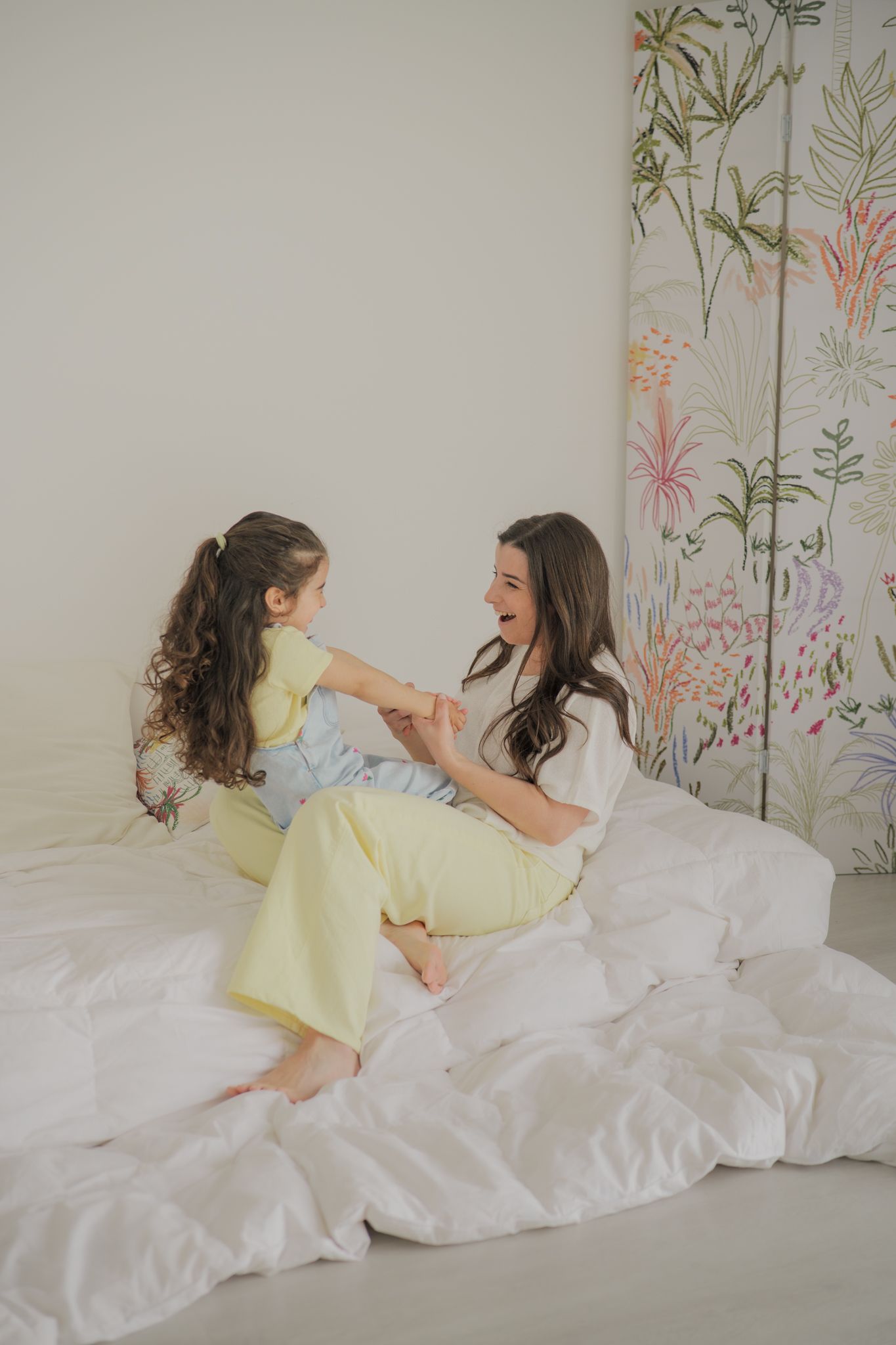A mother and daughter in matching yellow pajamas laugh together on a white bed near a floral room divider.
