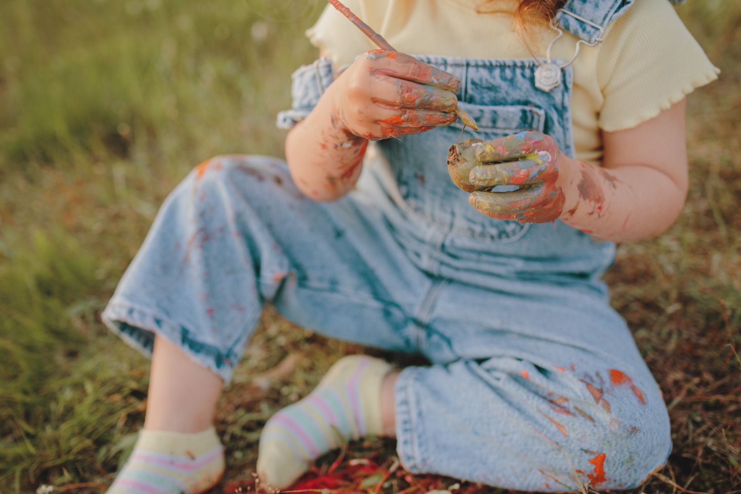 Child in blue overalls and white top sitting on grass with a book.