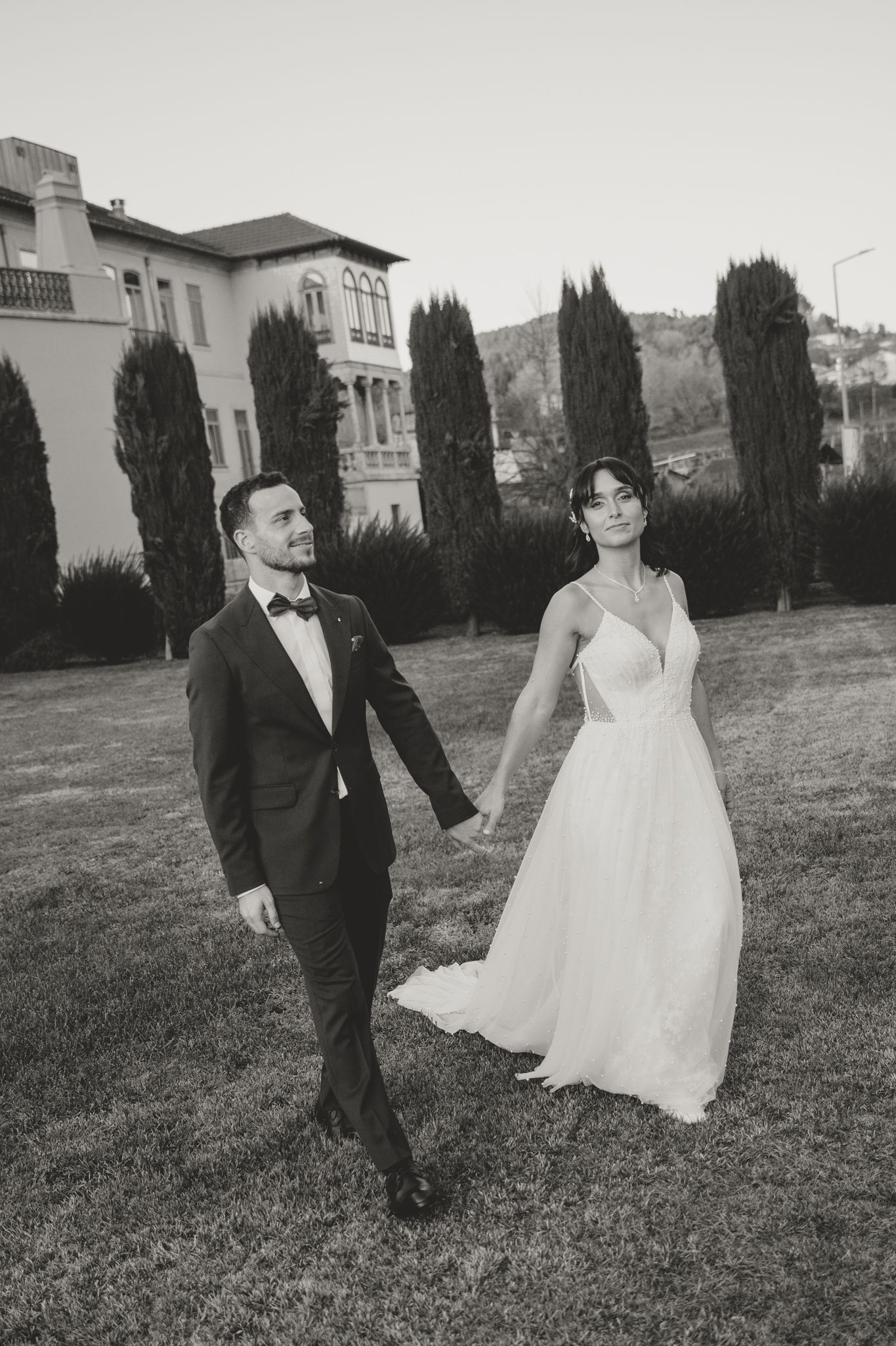 A couple in formal wedding attire walks together on a grassy lawn with buildings in the background.
