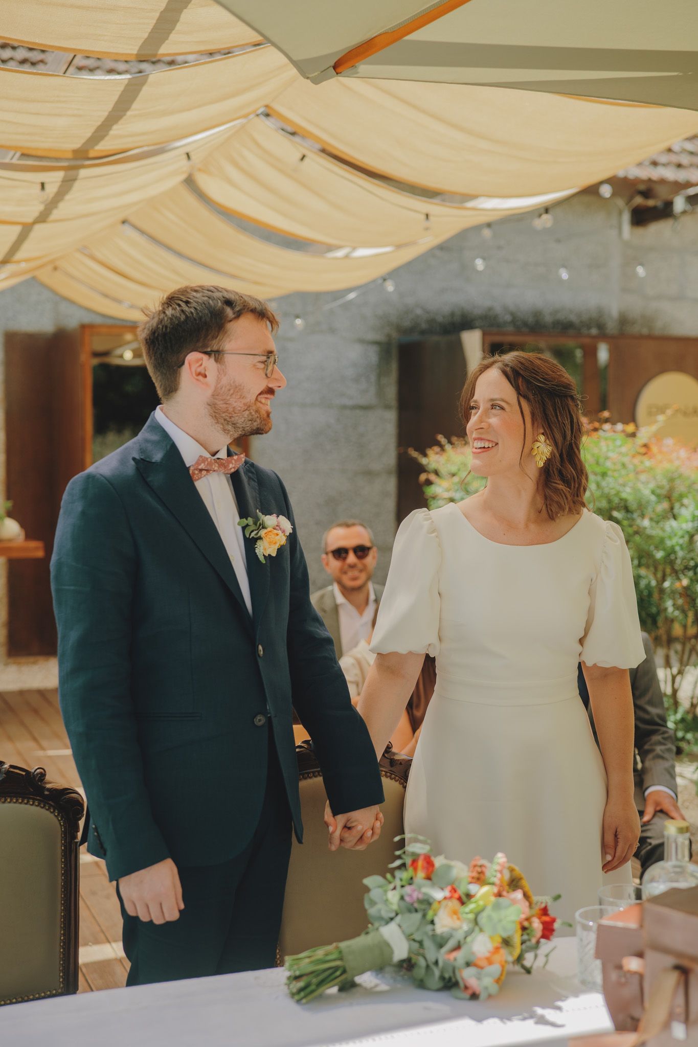 A couple shares a candid moment under a cream-colored canopy at an outdoor celebration.