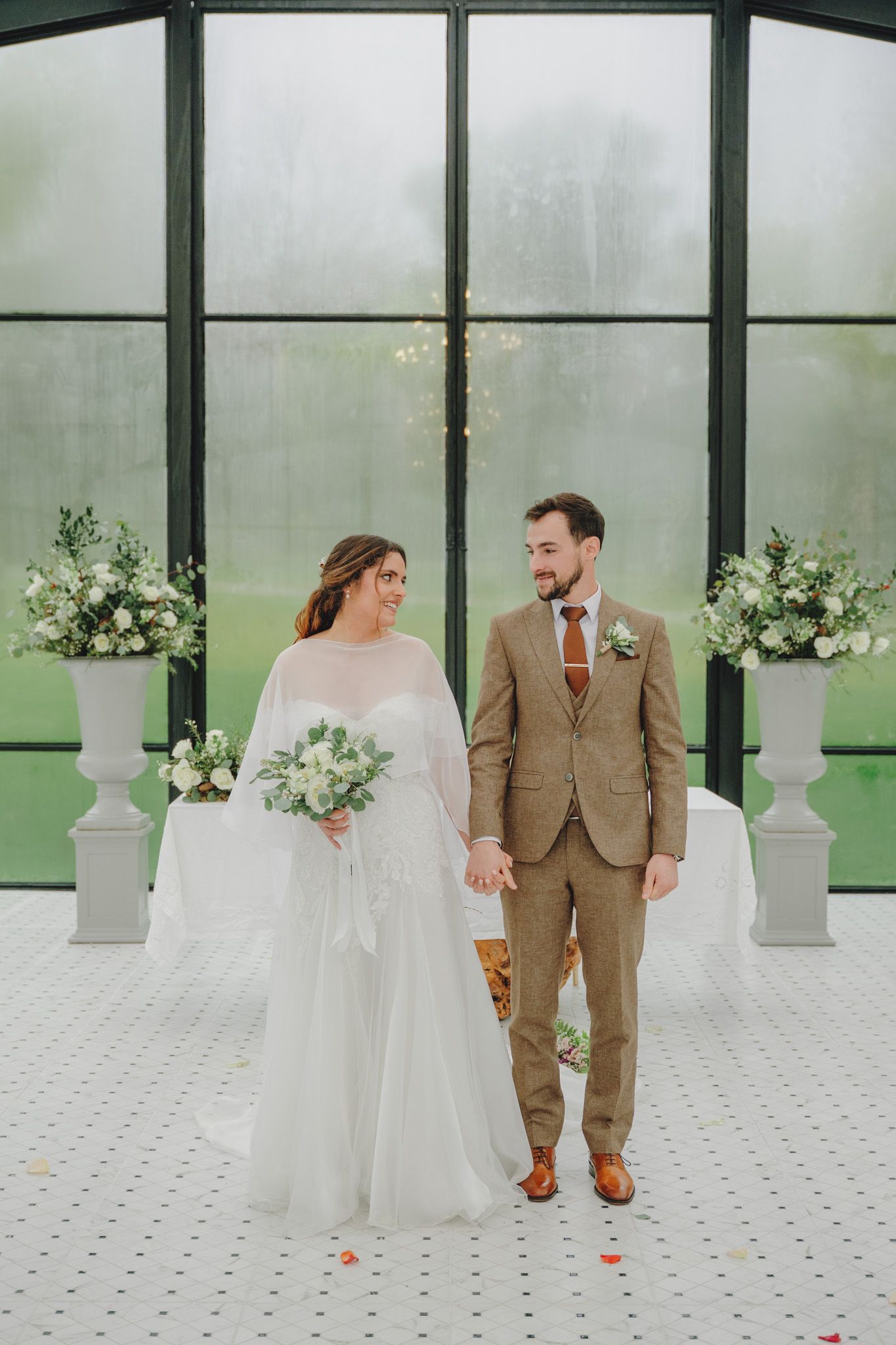Newlyweds walk hand-in-hand through modern greenhouse venue decorated with white flowers.