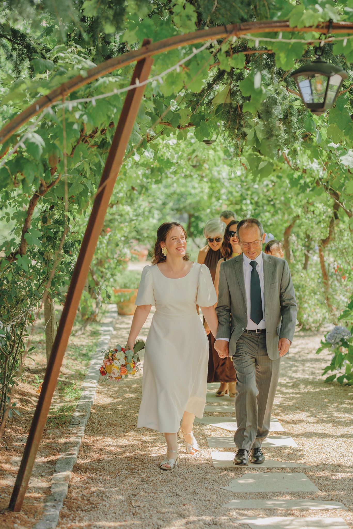 A couple walks together under a canopy of green grape vines in a vineyard on a sunny afternoon.