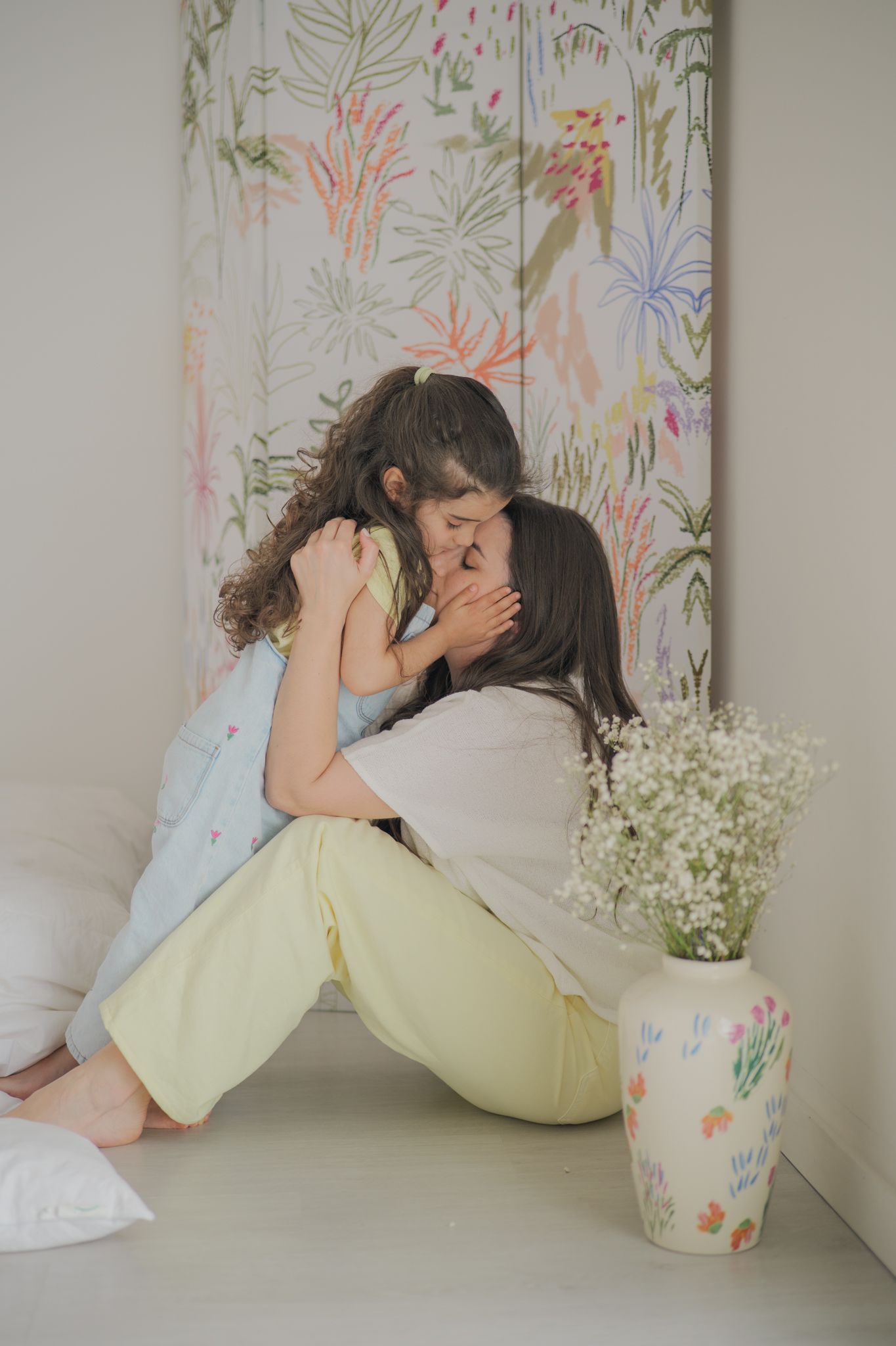 A mother and daughter share a tender embrace on a white floor beside a floral vase and botanical wall art.