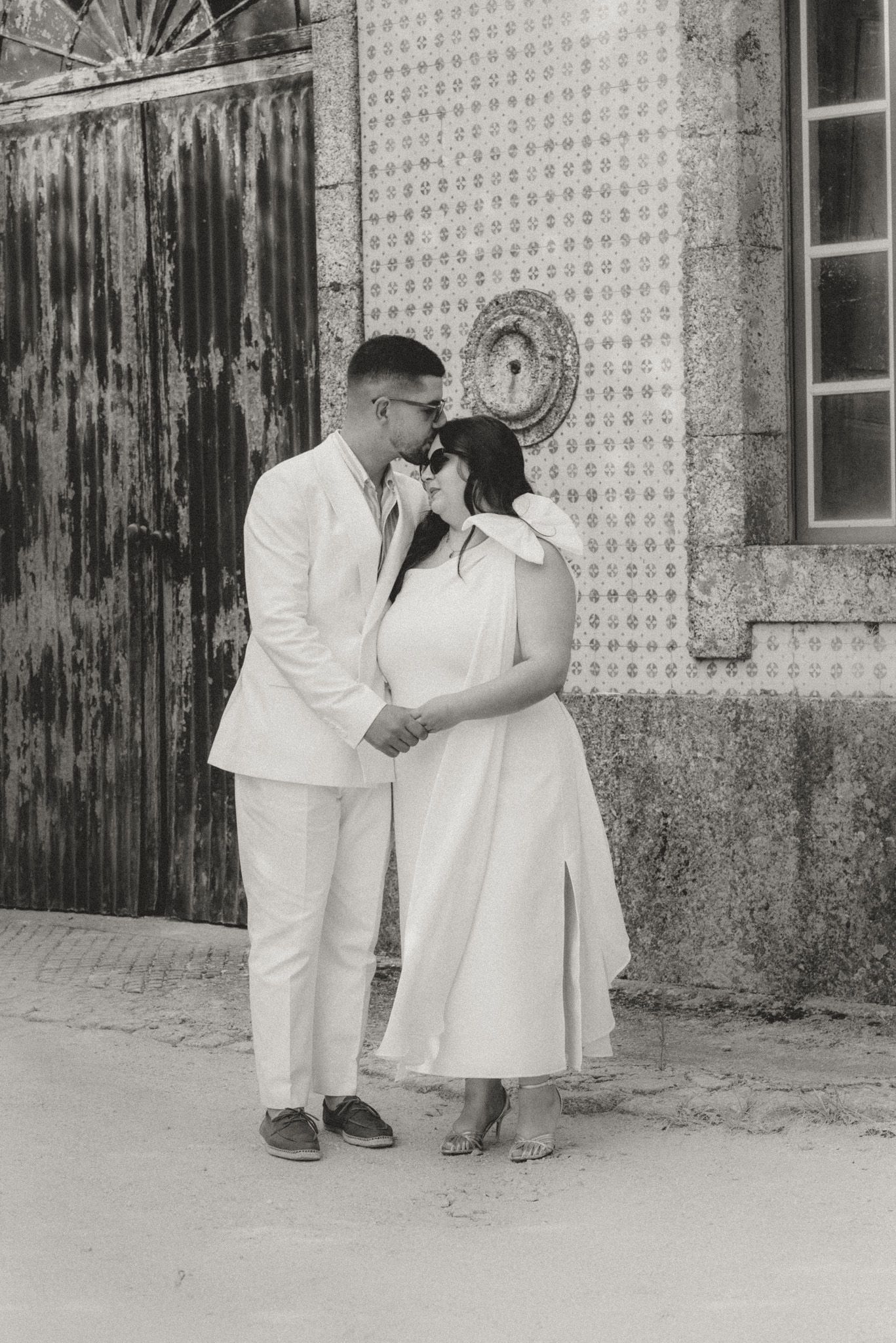 Two people in white outfits share an intimate moment against a textured wall.