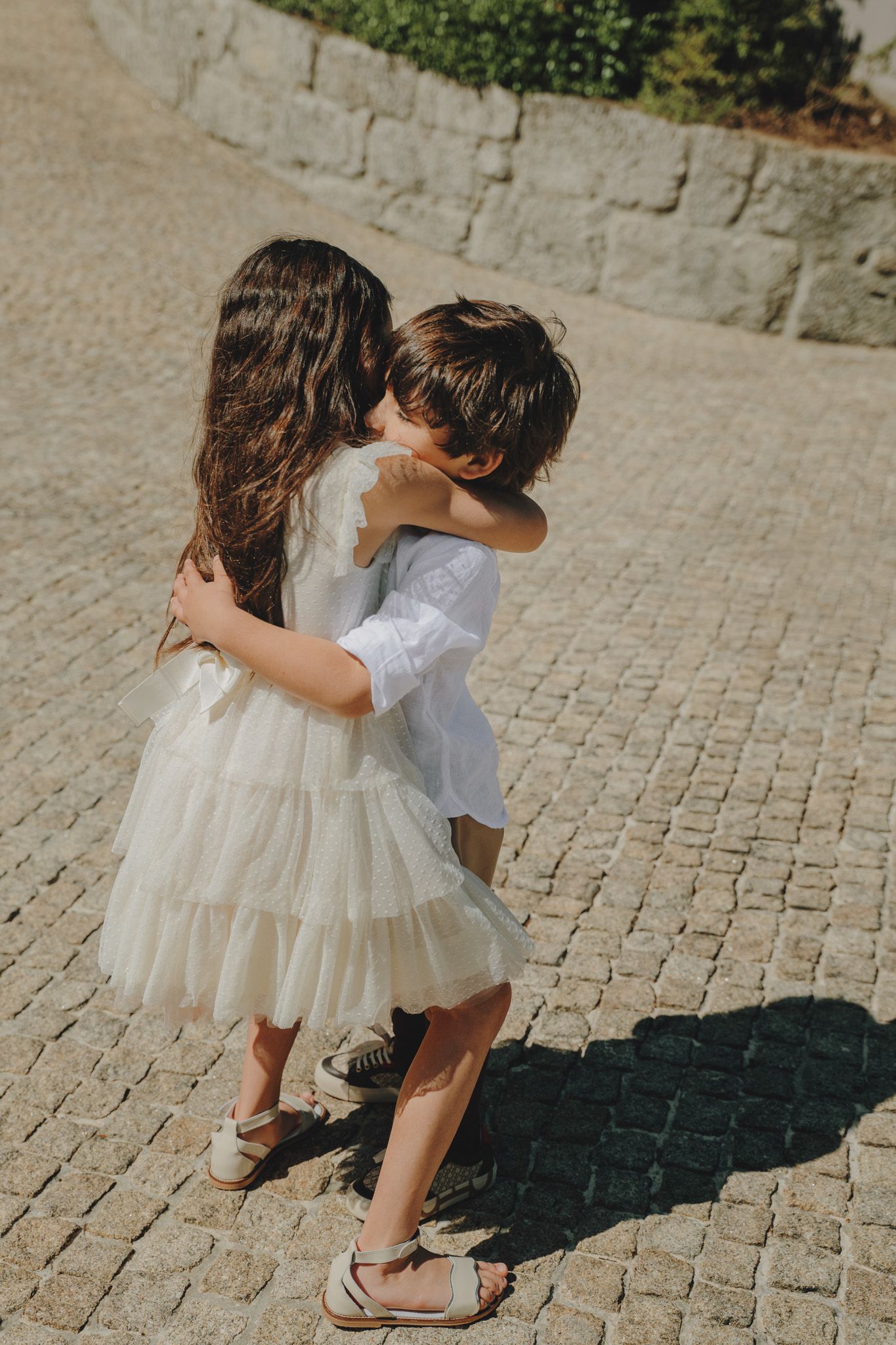 Two children in white outfits playing and hugging on cobblestone ground.