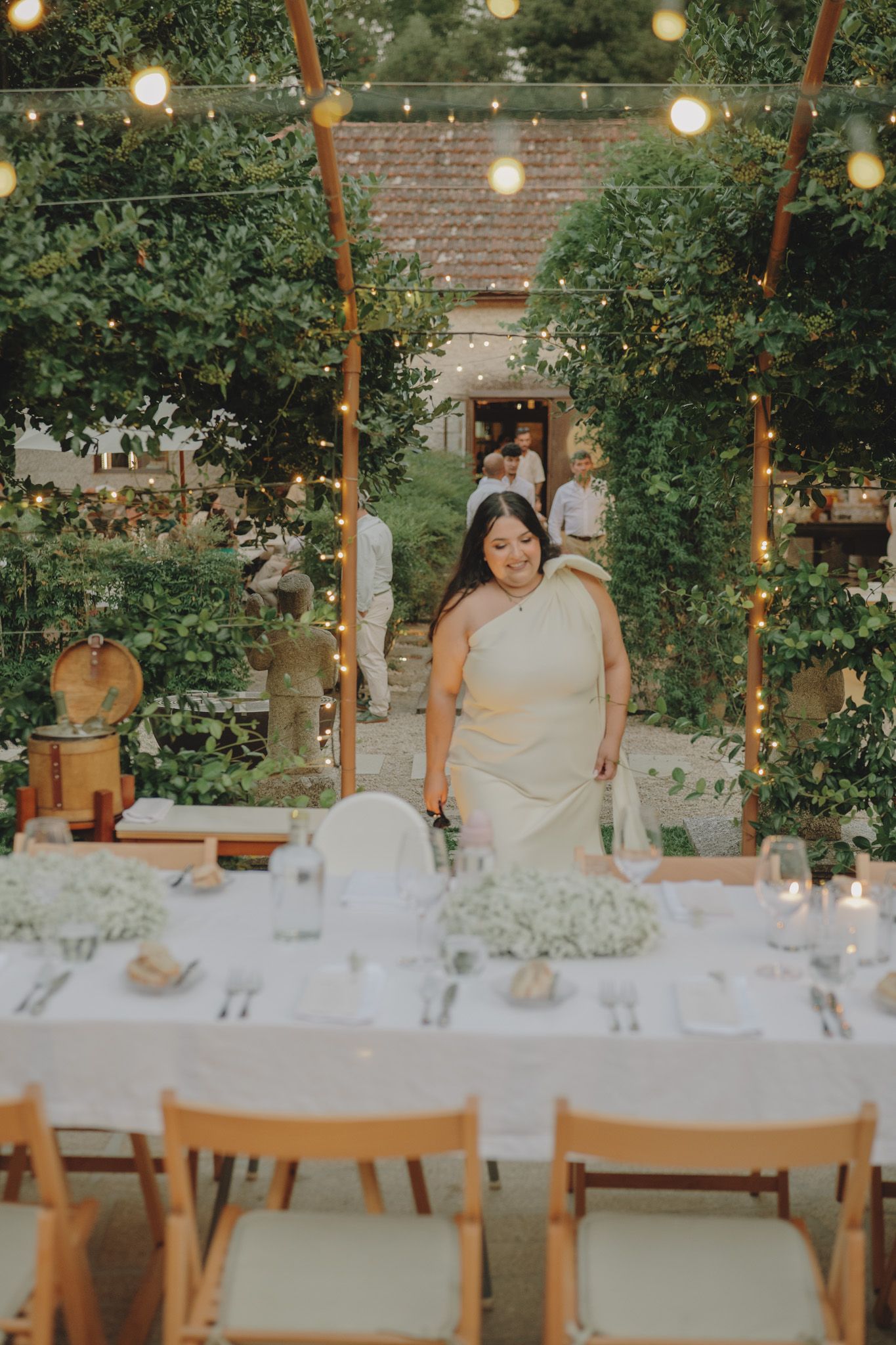 An outdoor wedding reception setup with string lights and elegant table settings among green foliage.