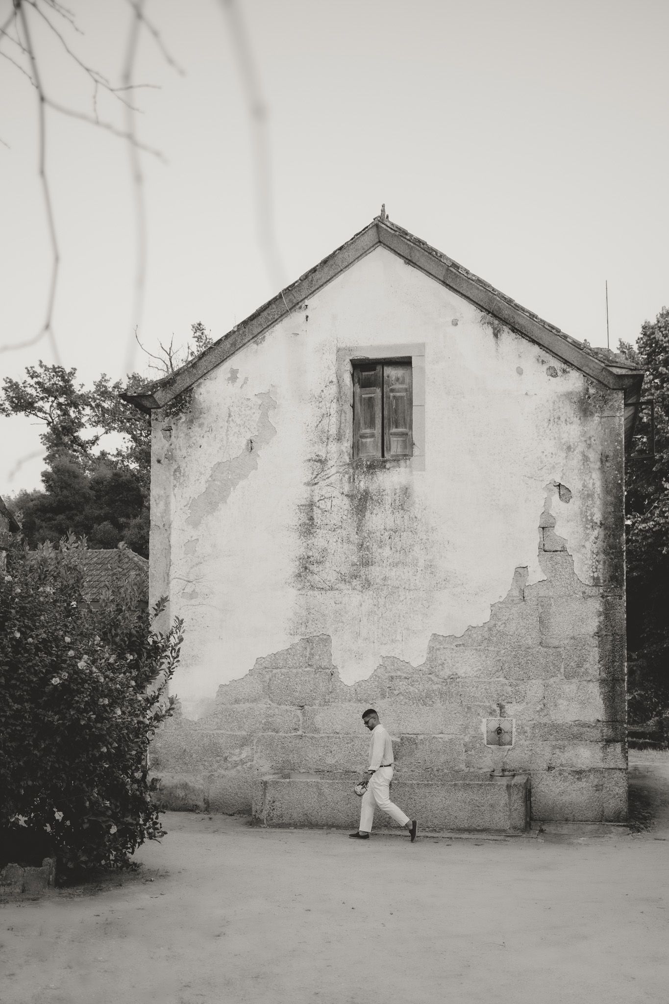 Black and white photograph of a person walking past a weathered white building wall with peeling paint and small windows.