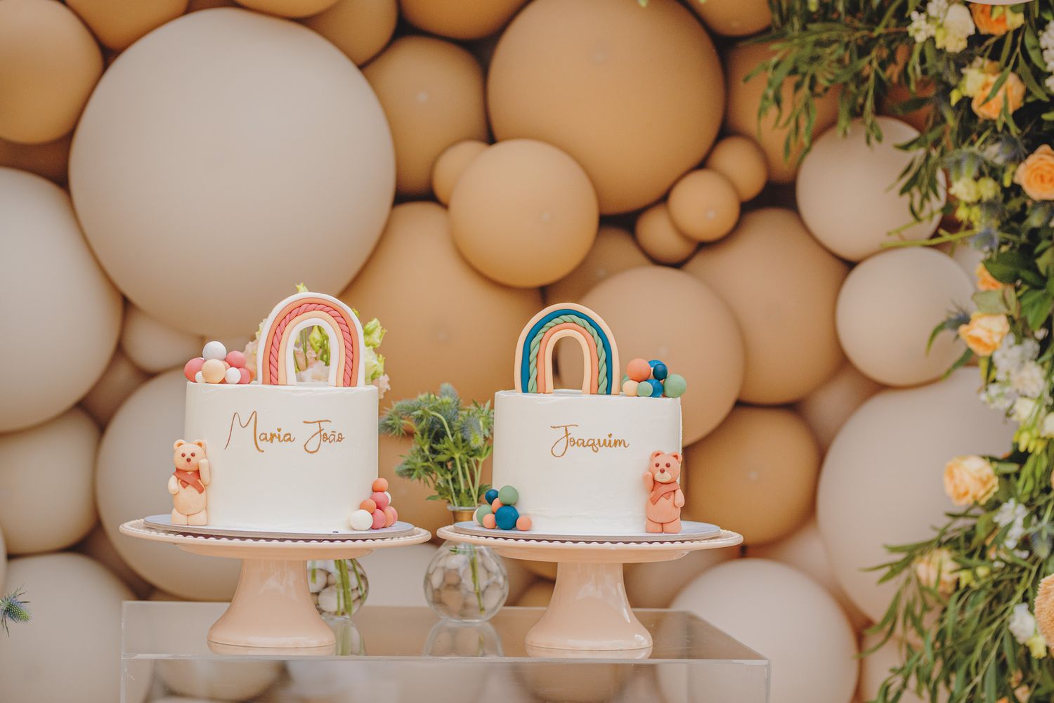 Two white celebration cakes with rainbow decorations displayed against a neutral balloon backdrop.