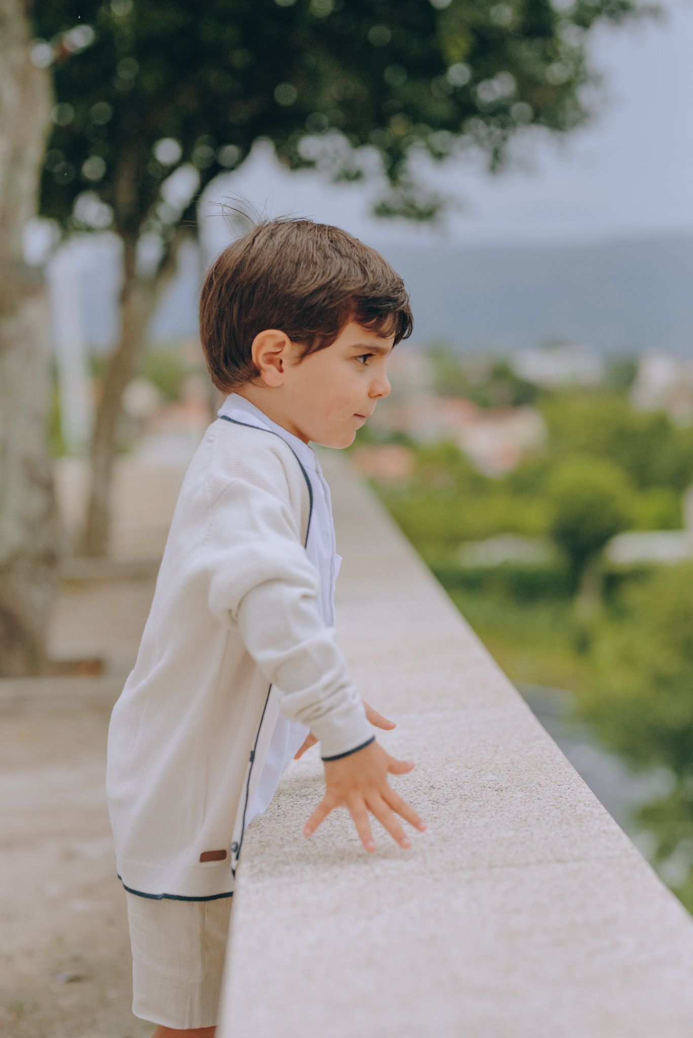 Child in white clothing leaning over a stone wall overlooking a scenic view.