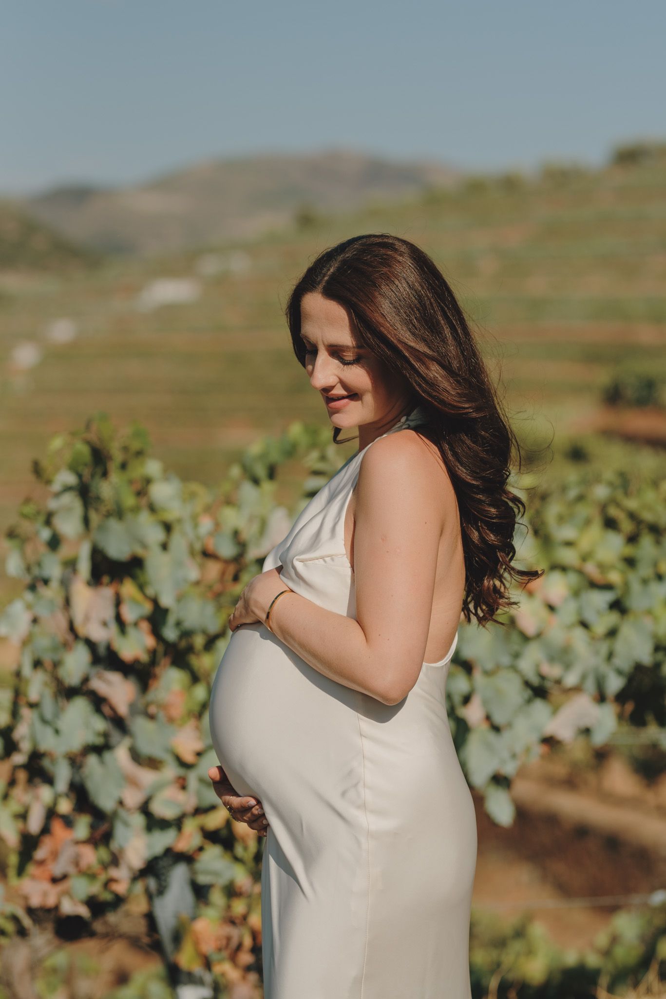 Woman in white dress poses in a sunny vineyard against rolling hills and blue sky.