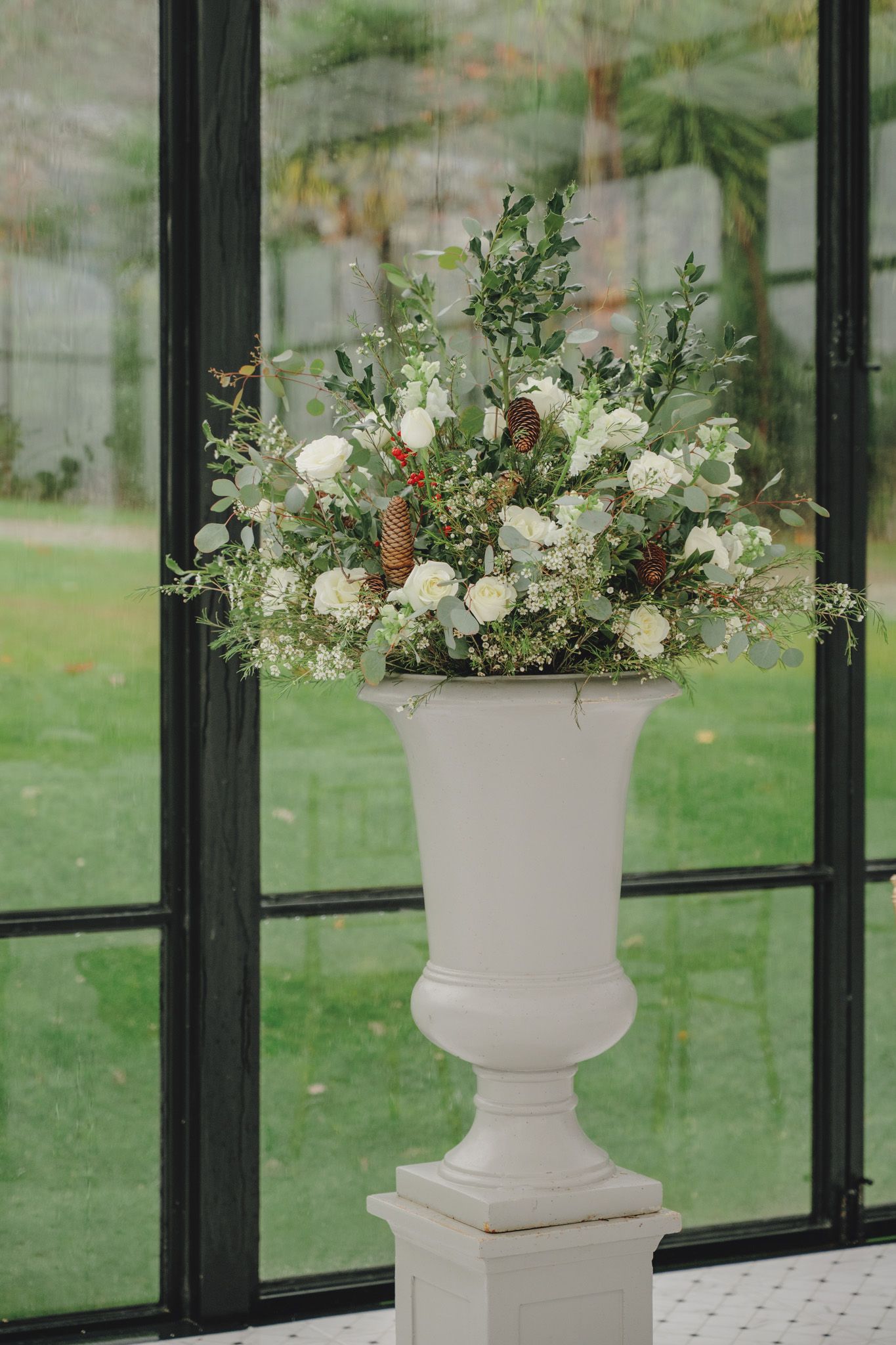 White floral arrangements in classic urns displayed against glass windows overlooking green lawn.