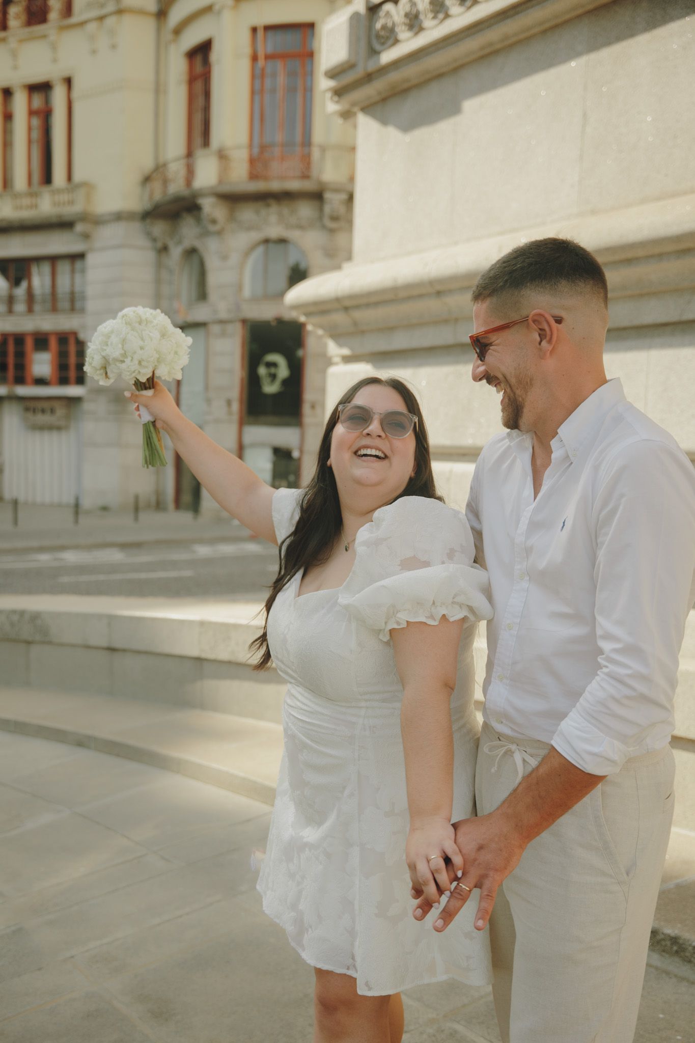 A couple in all-white outfits poses together against a classic architectural backdrop.