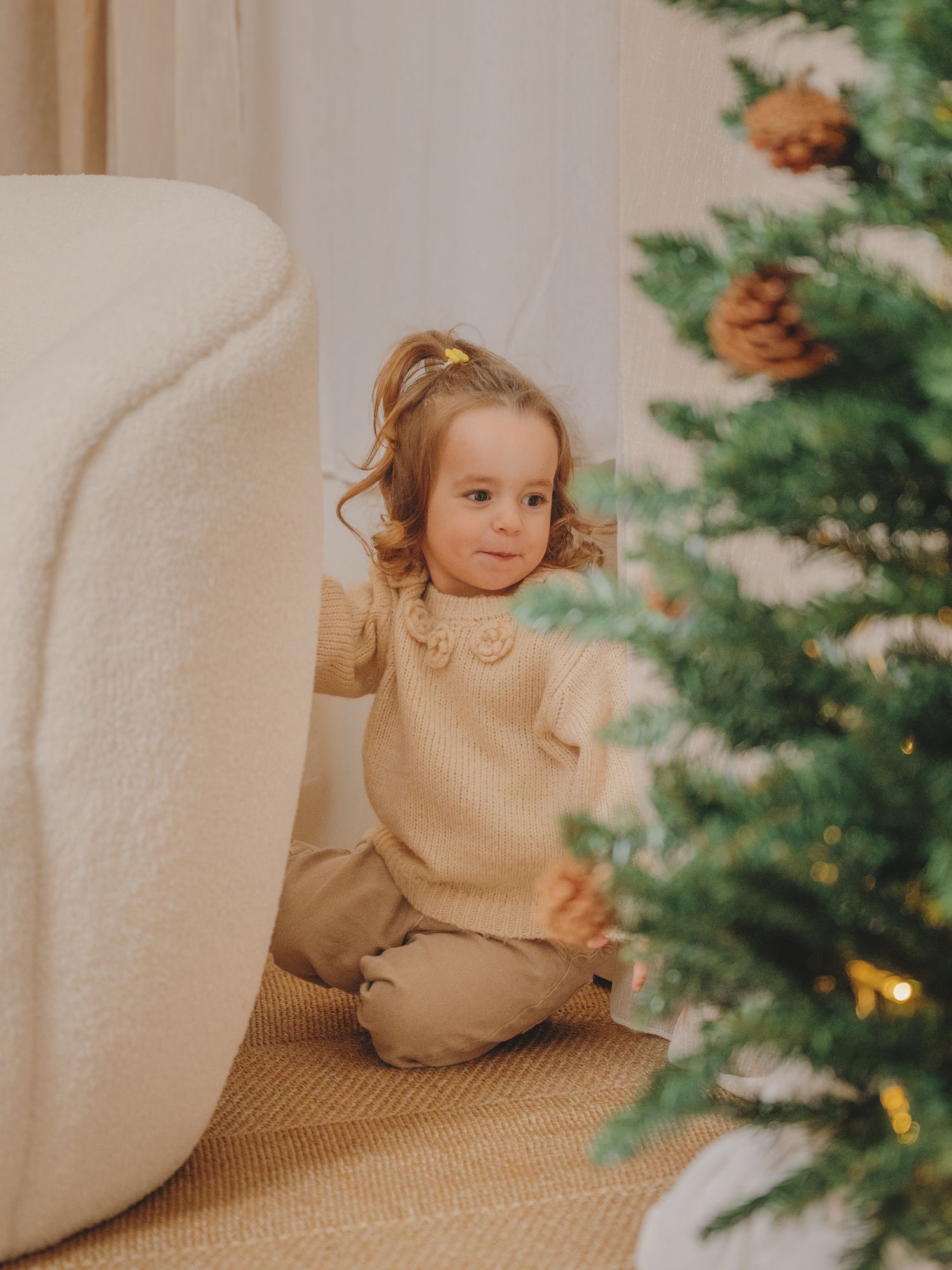 Toddler in a cream knit sweater and beige pants kneels beside a cozy boucle chair near a decorated Christmas tree with pinecones.