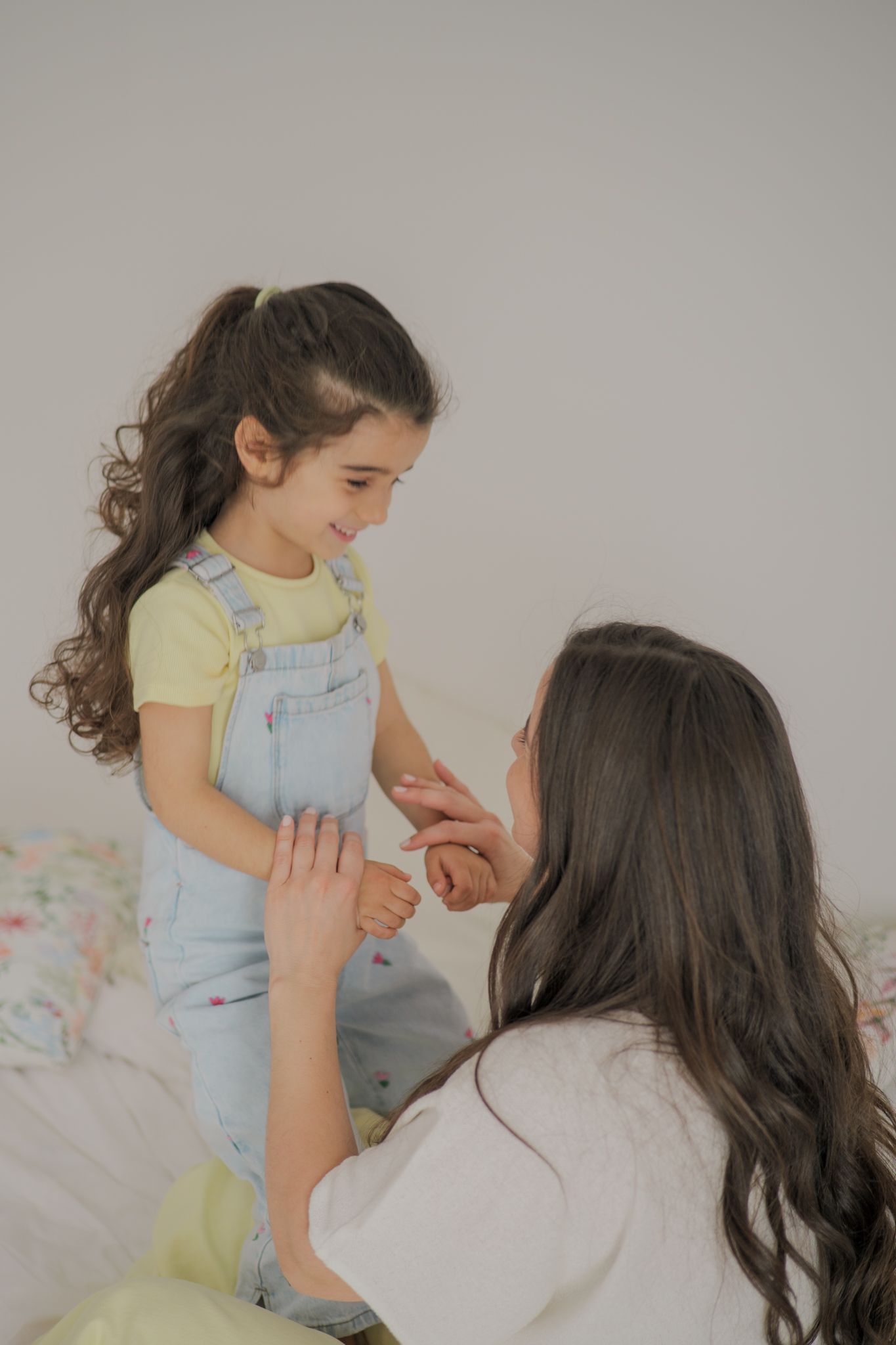 A mother kneeling on a bed, holding hands with her smiling young daughter in denim overalls.