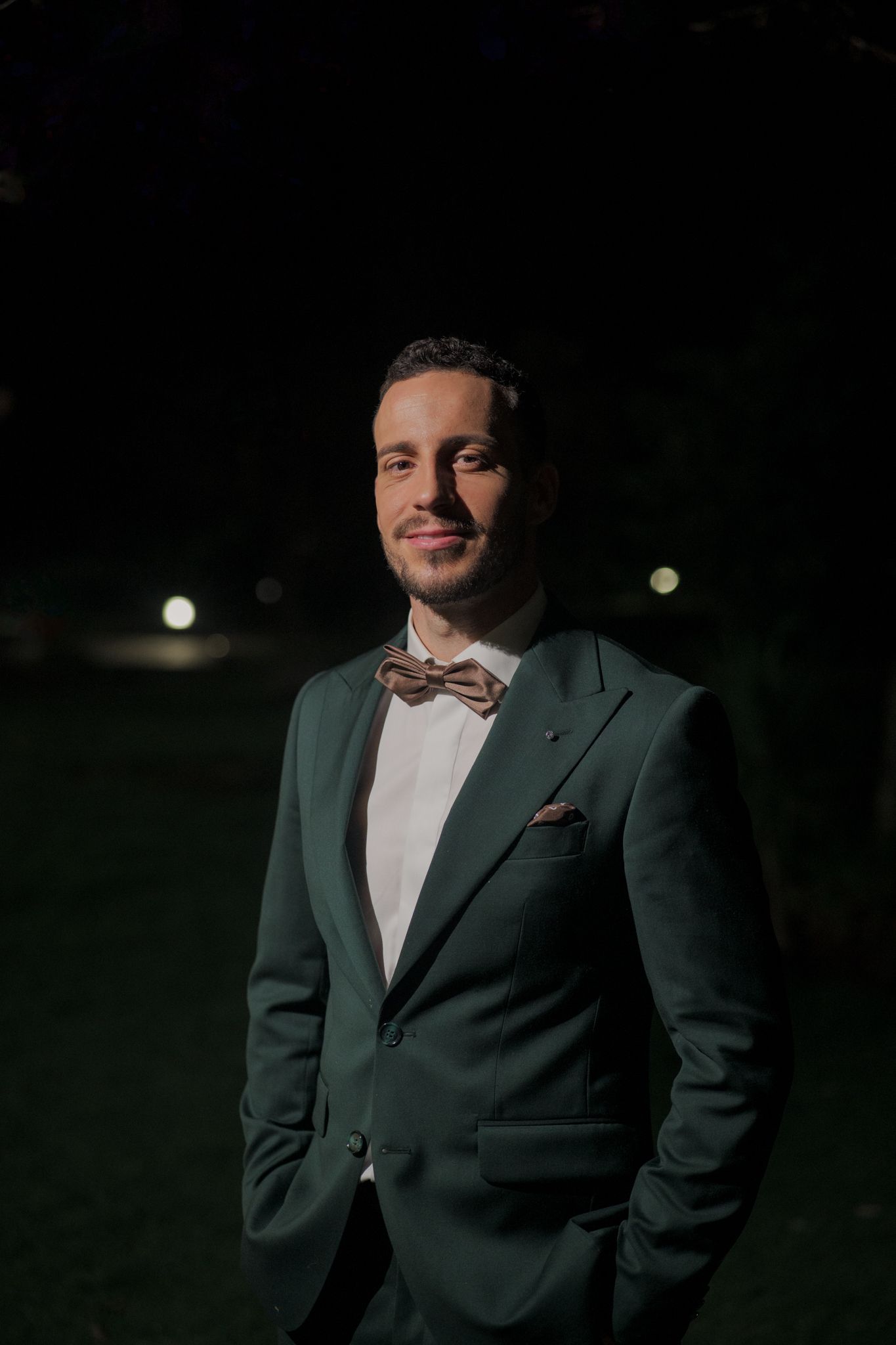 A person in a dark green suit and bowtie poses in dramatic evening lighting.
