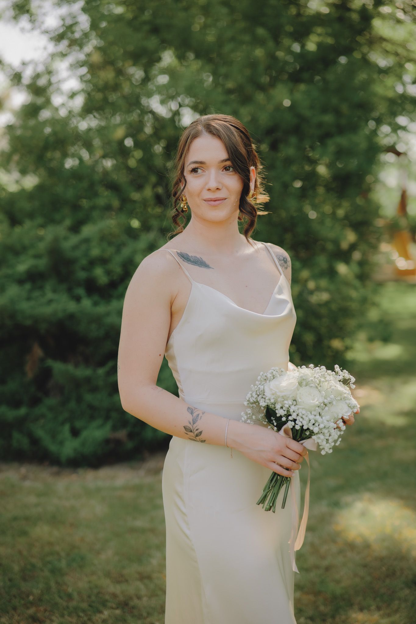 Elegant bride in simple white slip dress holds classic white bouquet in outdoor garden setting.