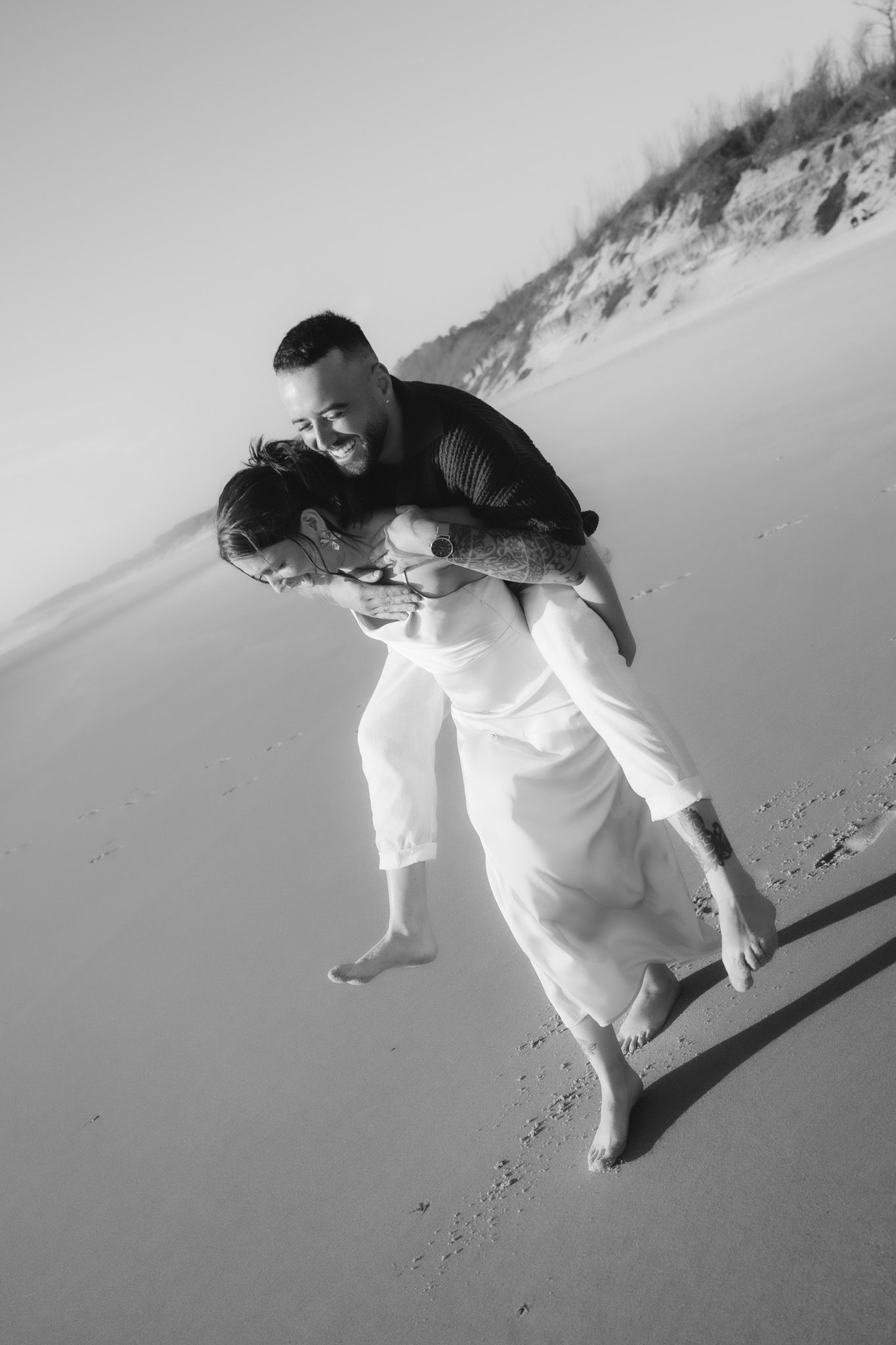 Playful couple in white clothing running and dancing on sandy beach.