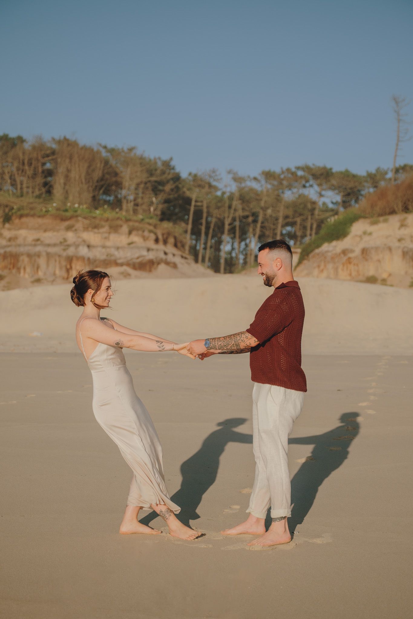 Two people dancing playfully on a sandy beach with pine trees in the background.