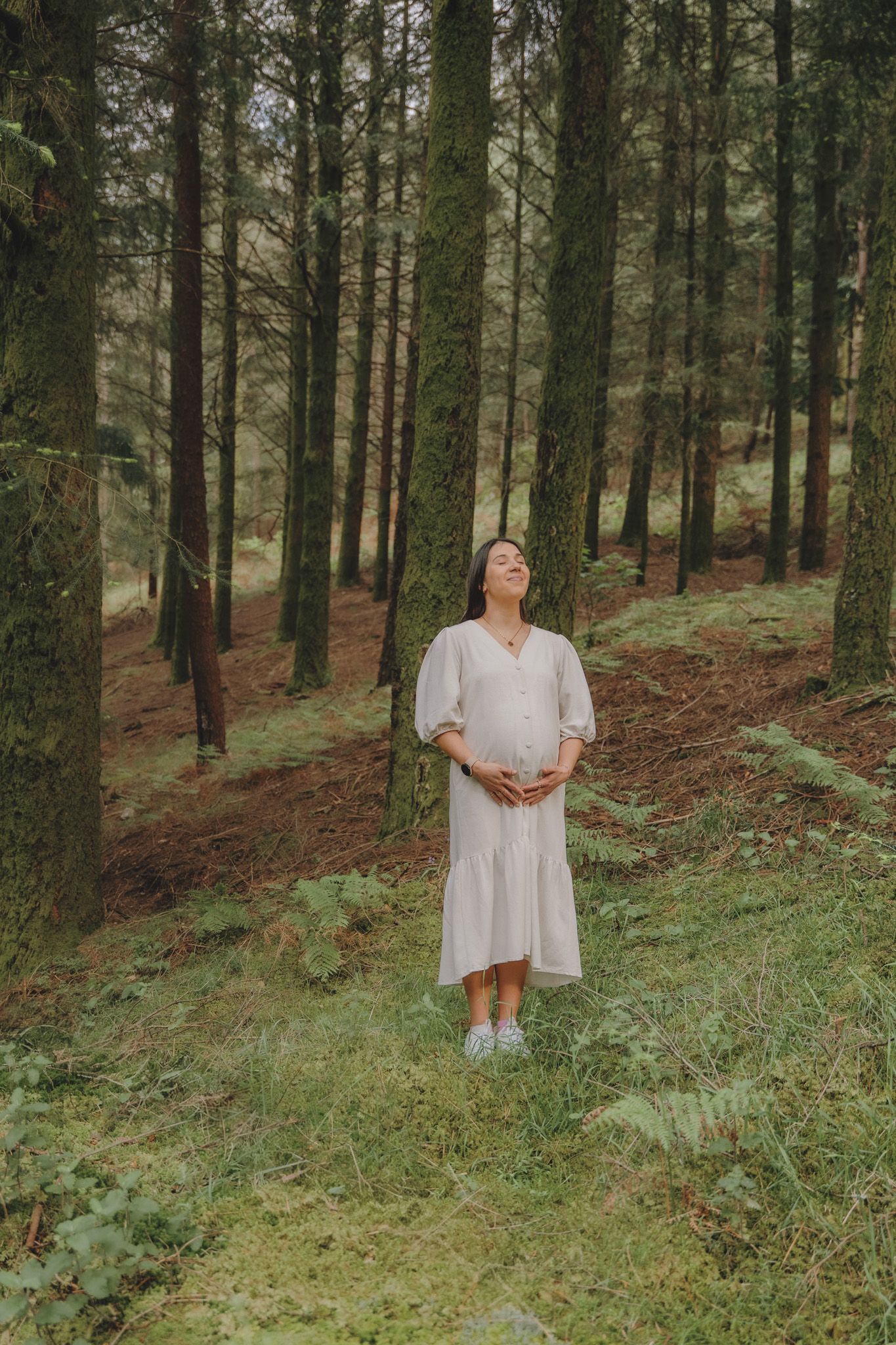 A person in a white dress stands among tall pine trees in a forest setting.