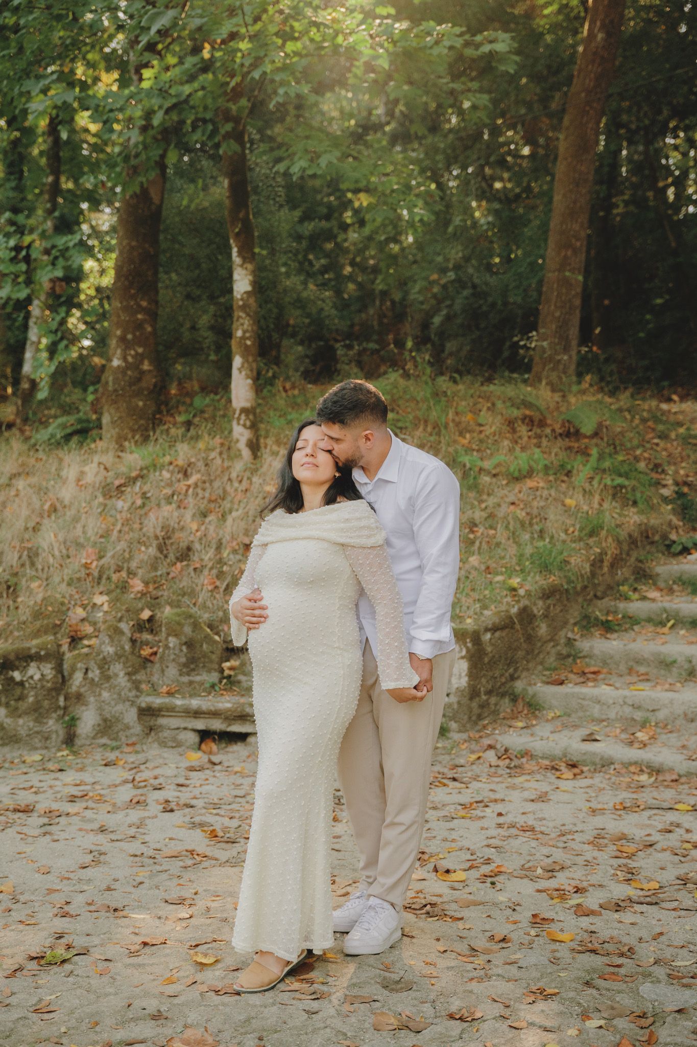 A tender maternity portrait against a wooded backdrop with fallen leaves.