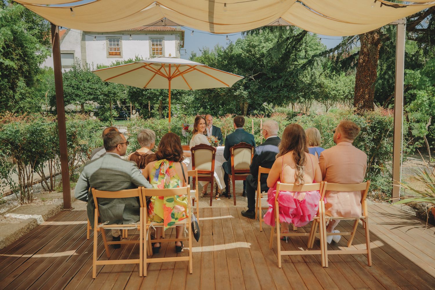 Guests seated under a beige canopy on a wooden deck for an outdoor wedding ceremony.