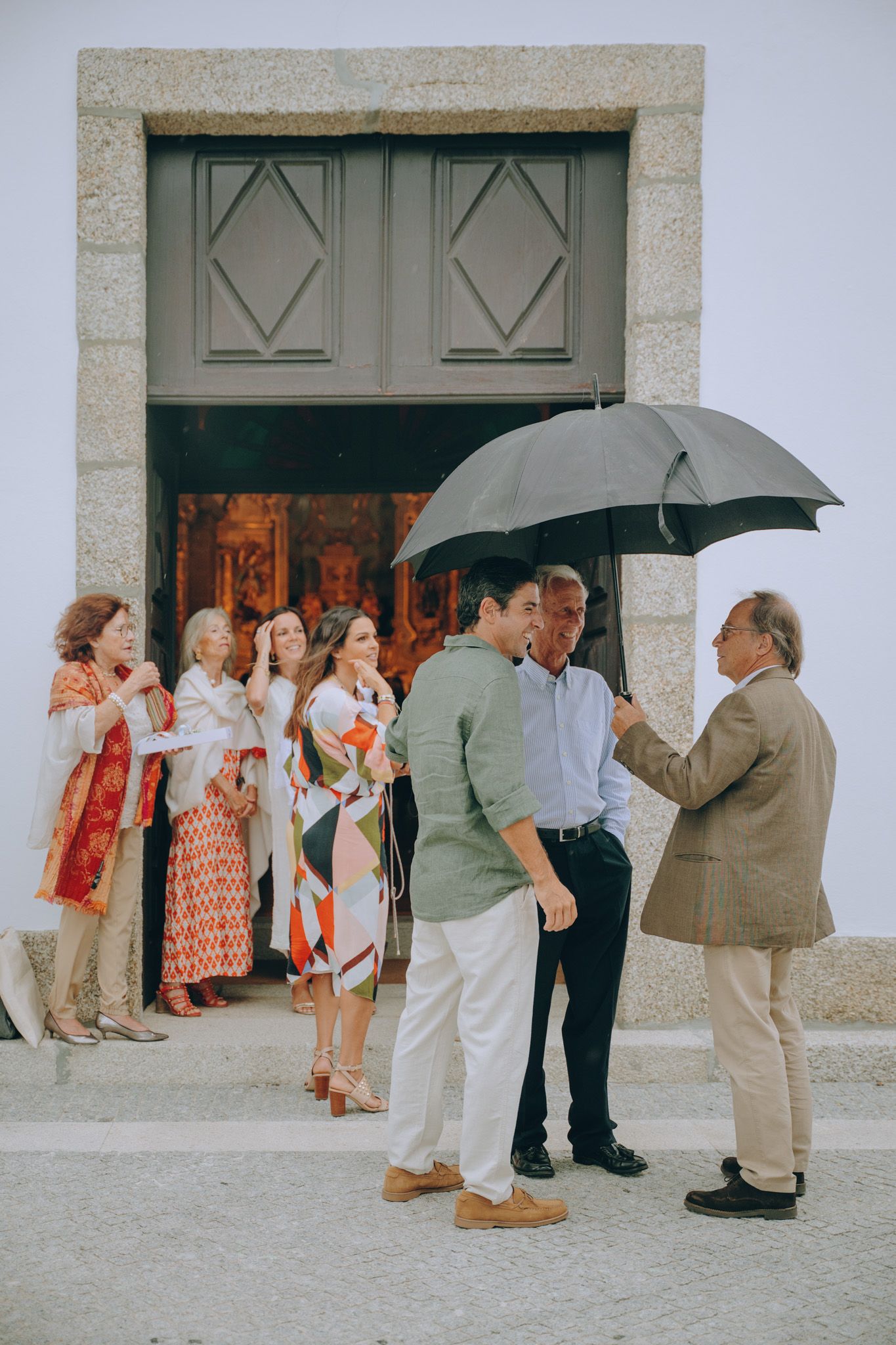 People wait in line under an umbrella outside a building entrance with ornate doors.