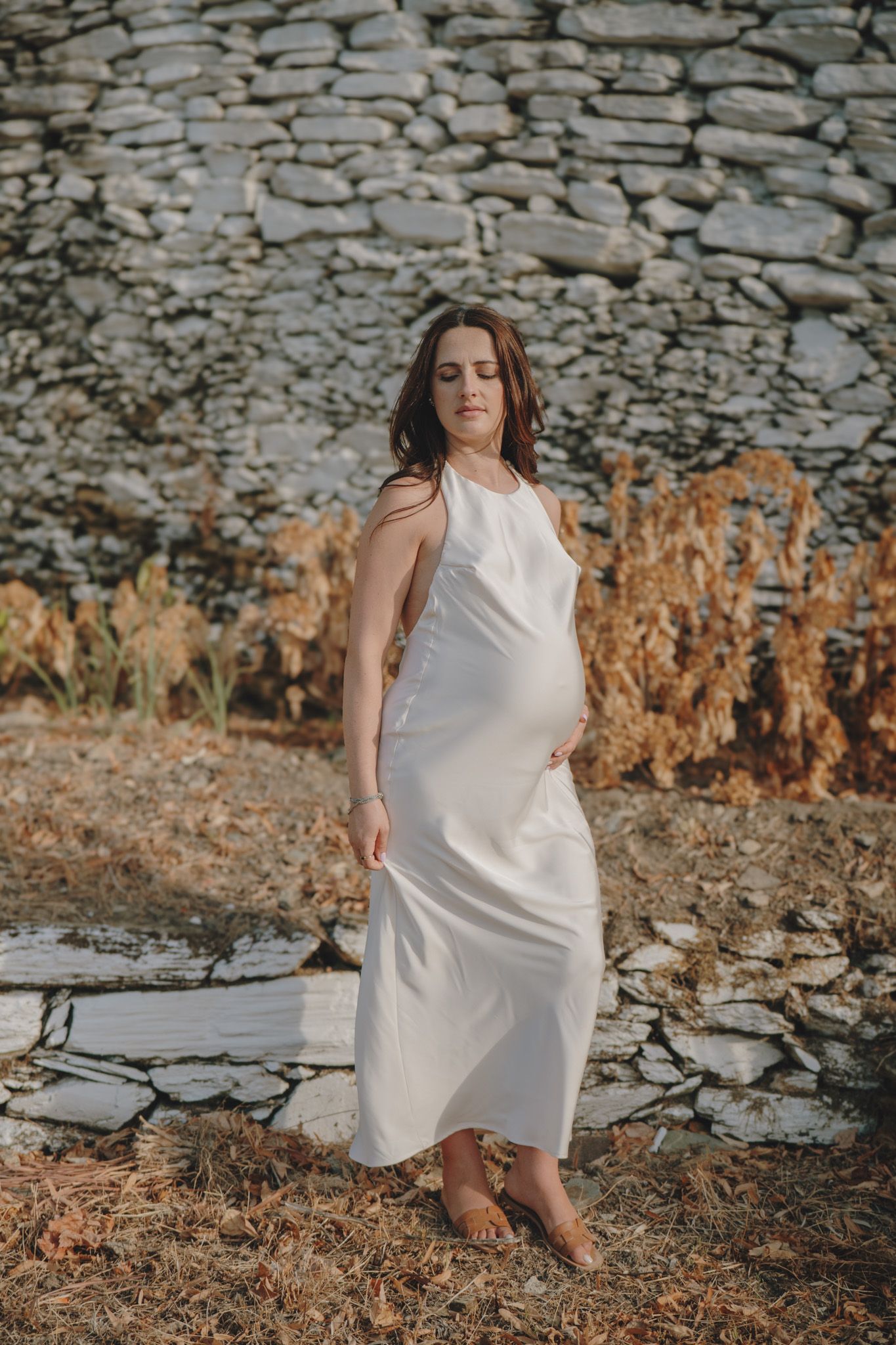 A figure in a white dress moves gracefully near an old stone wall in warm sunlight.