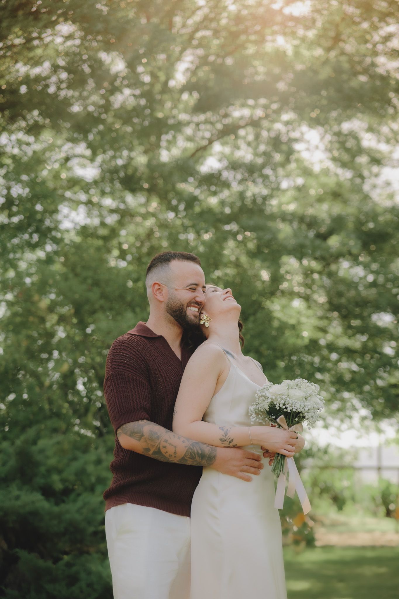 A couple embraces and laughs while holding a white bouquet in a sunny garden setting.