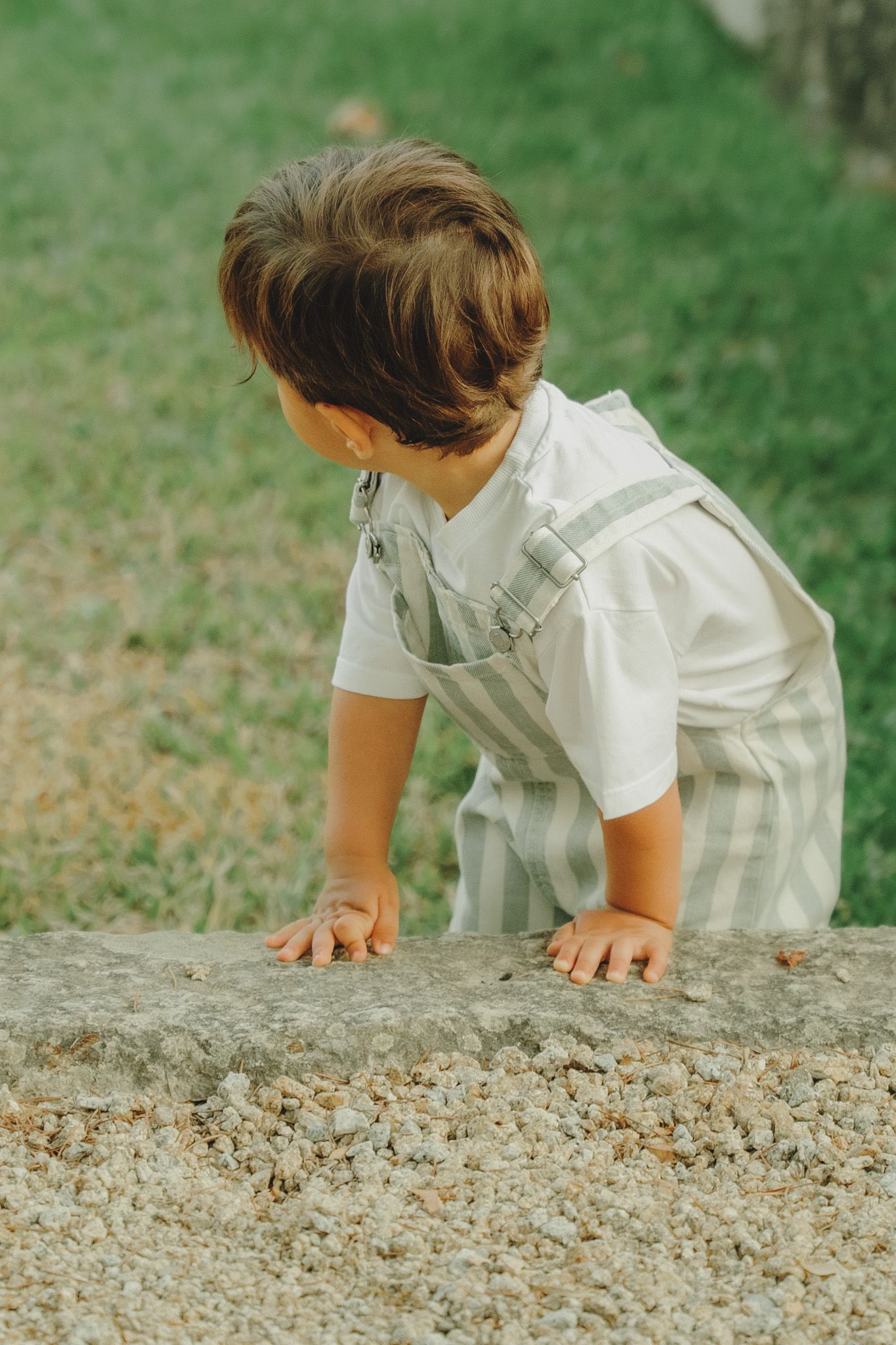 A child in white clothing leans over a stone wall, exploring with their hands in a curious pose.