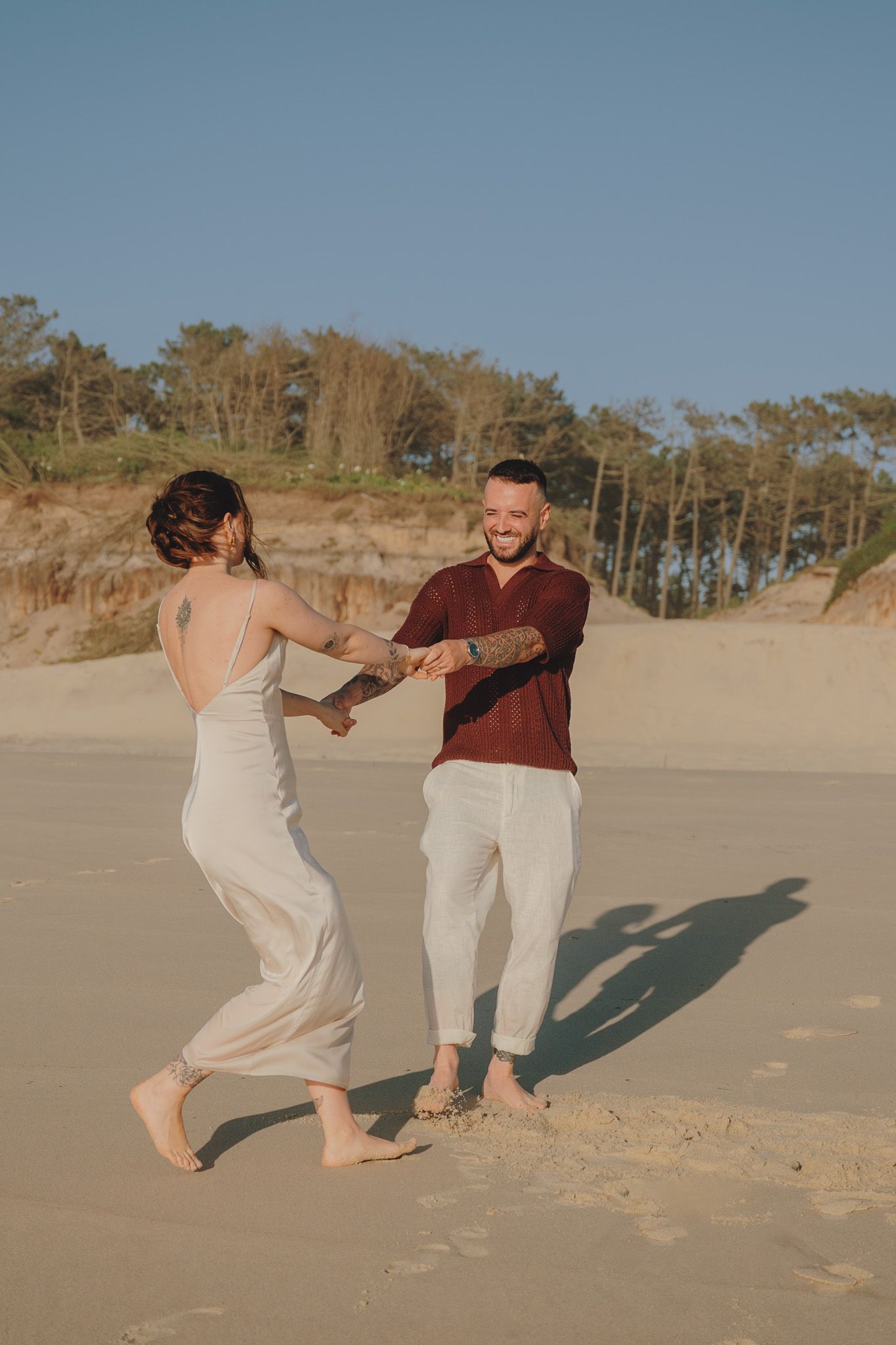 Couple in coordinated outfits dancing barefoot on a sunny beach.