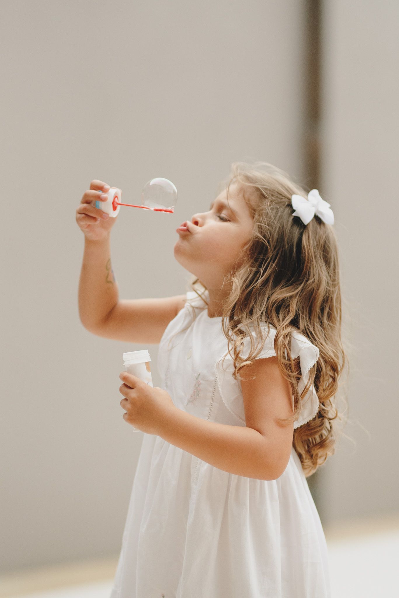 Young child in white dress blowing bubbles with joyful expression.