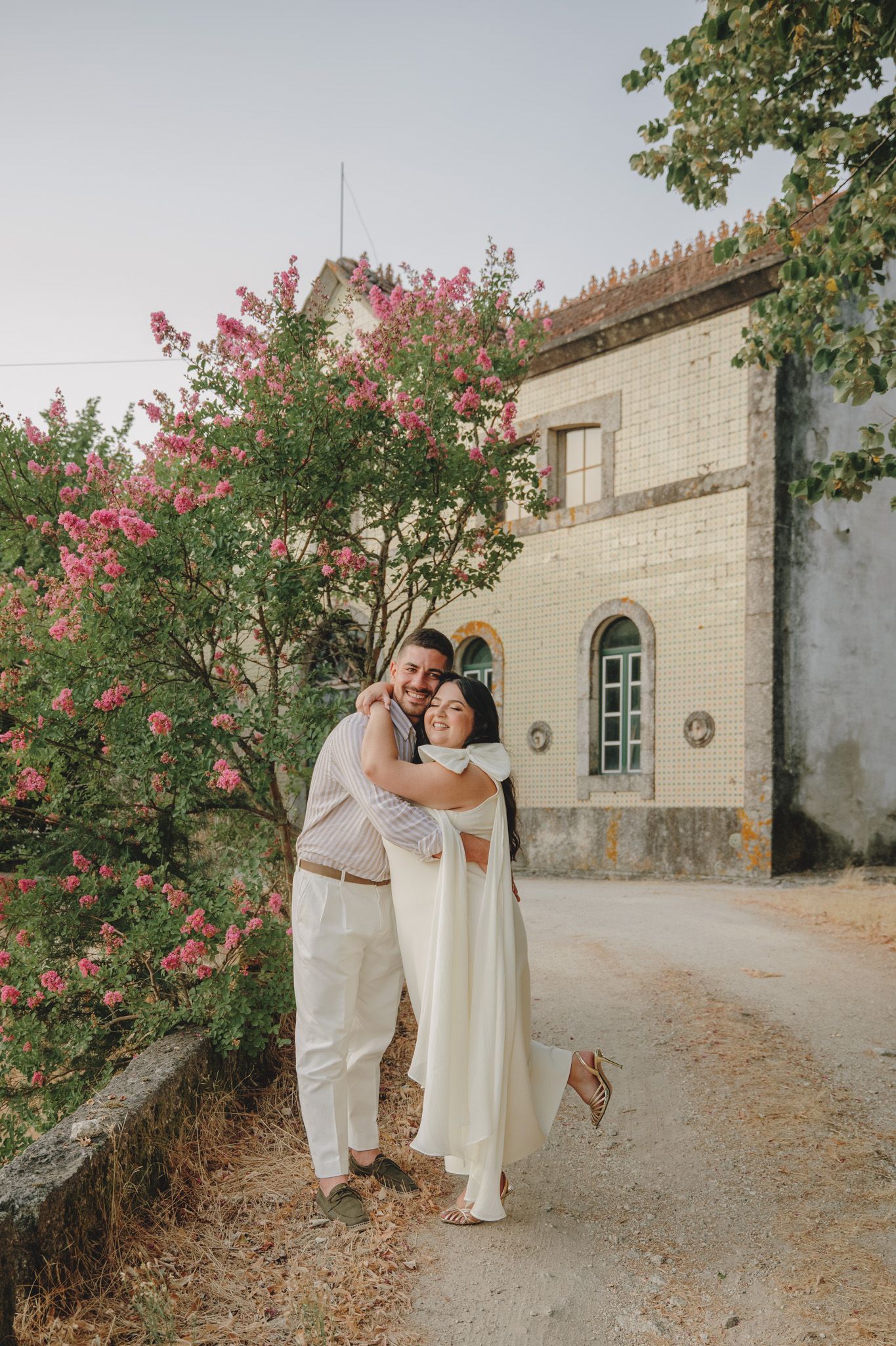 A couple in elegant white attire poses romantically against a stone building with pink flowering trees.