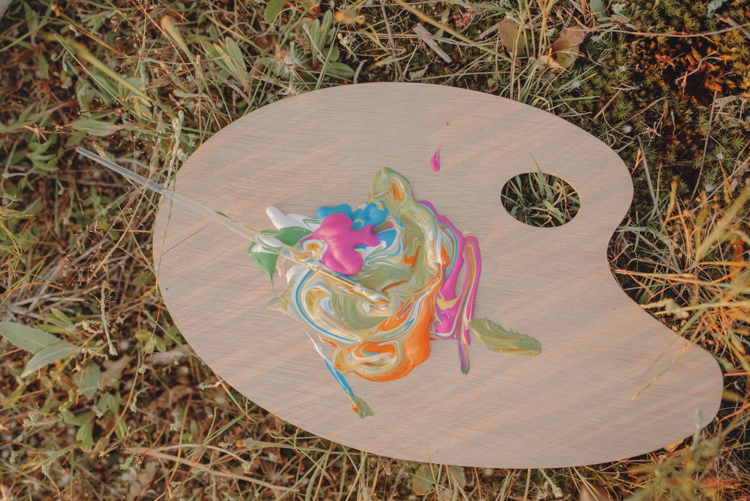 Colorful paint swirls decorate a wooden artist's palette against dried grass.