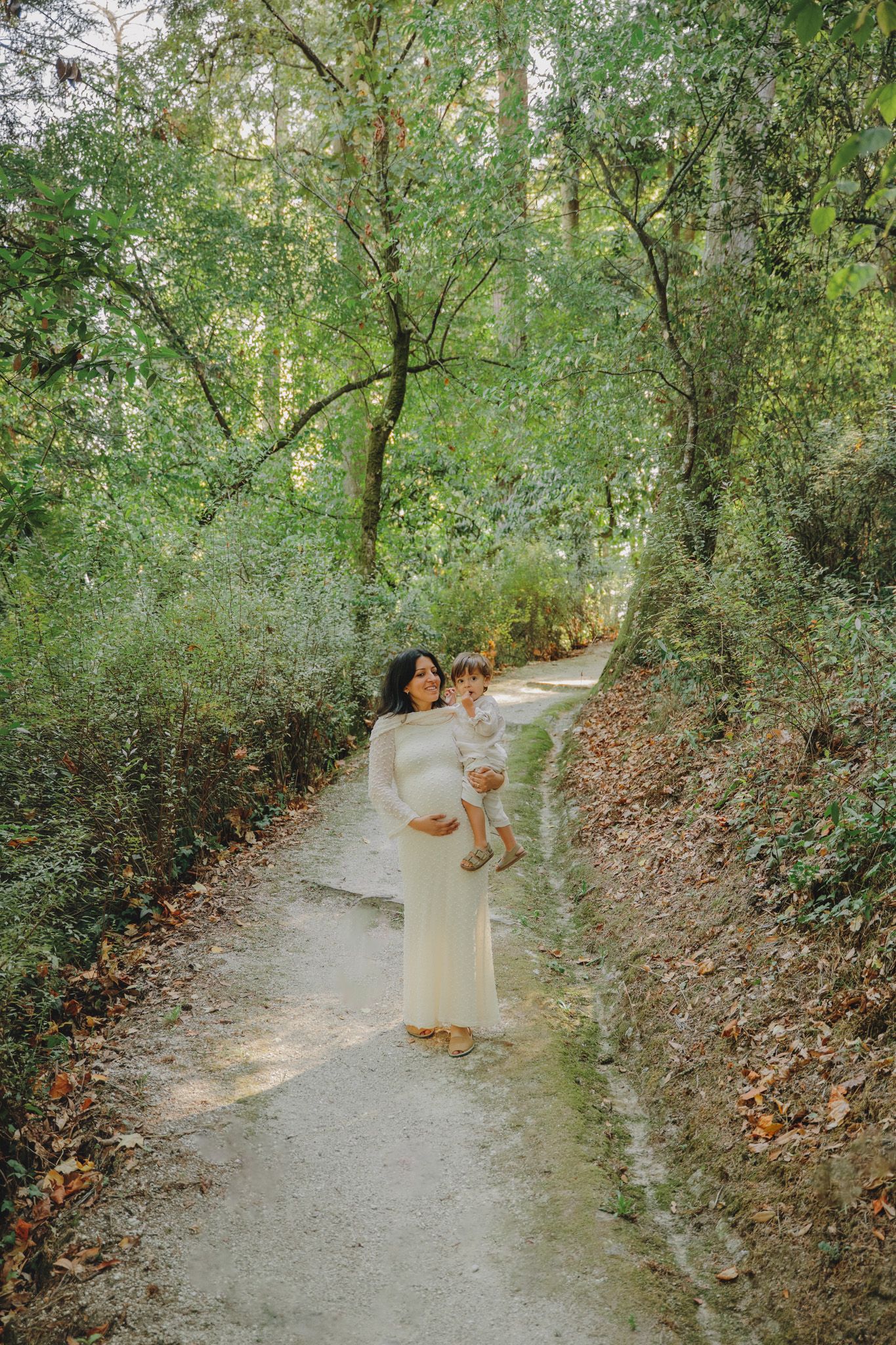 A woman in white holds a child while walking along a serene forest path.