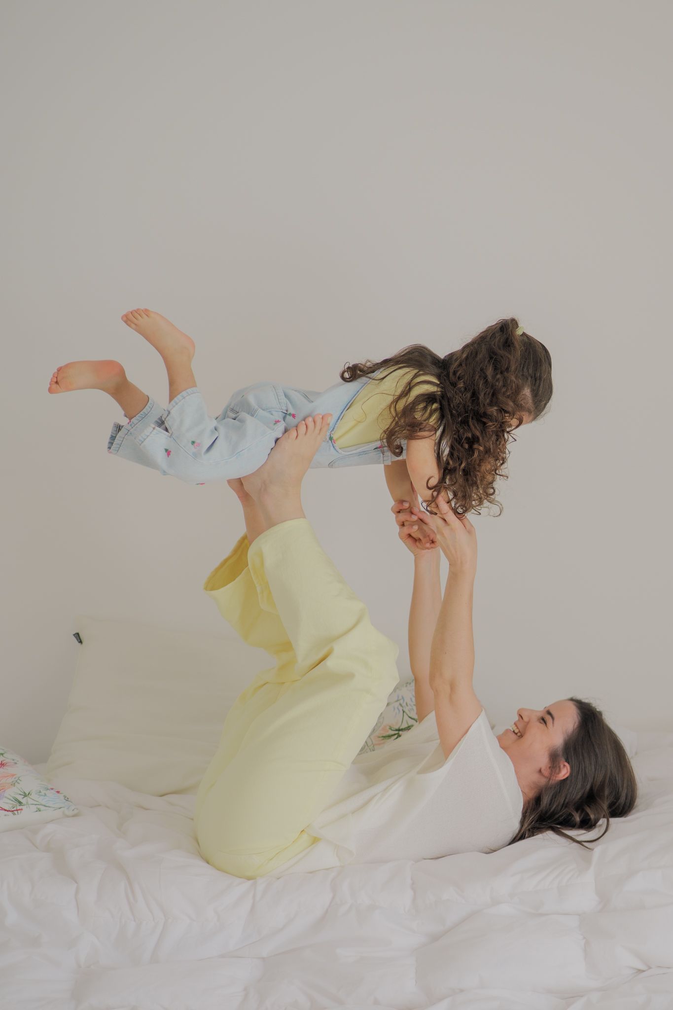 A mother lying on a bed lifts her young daughter playfully into the air, both wearing pastel pajamas.