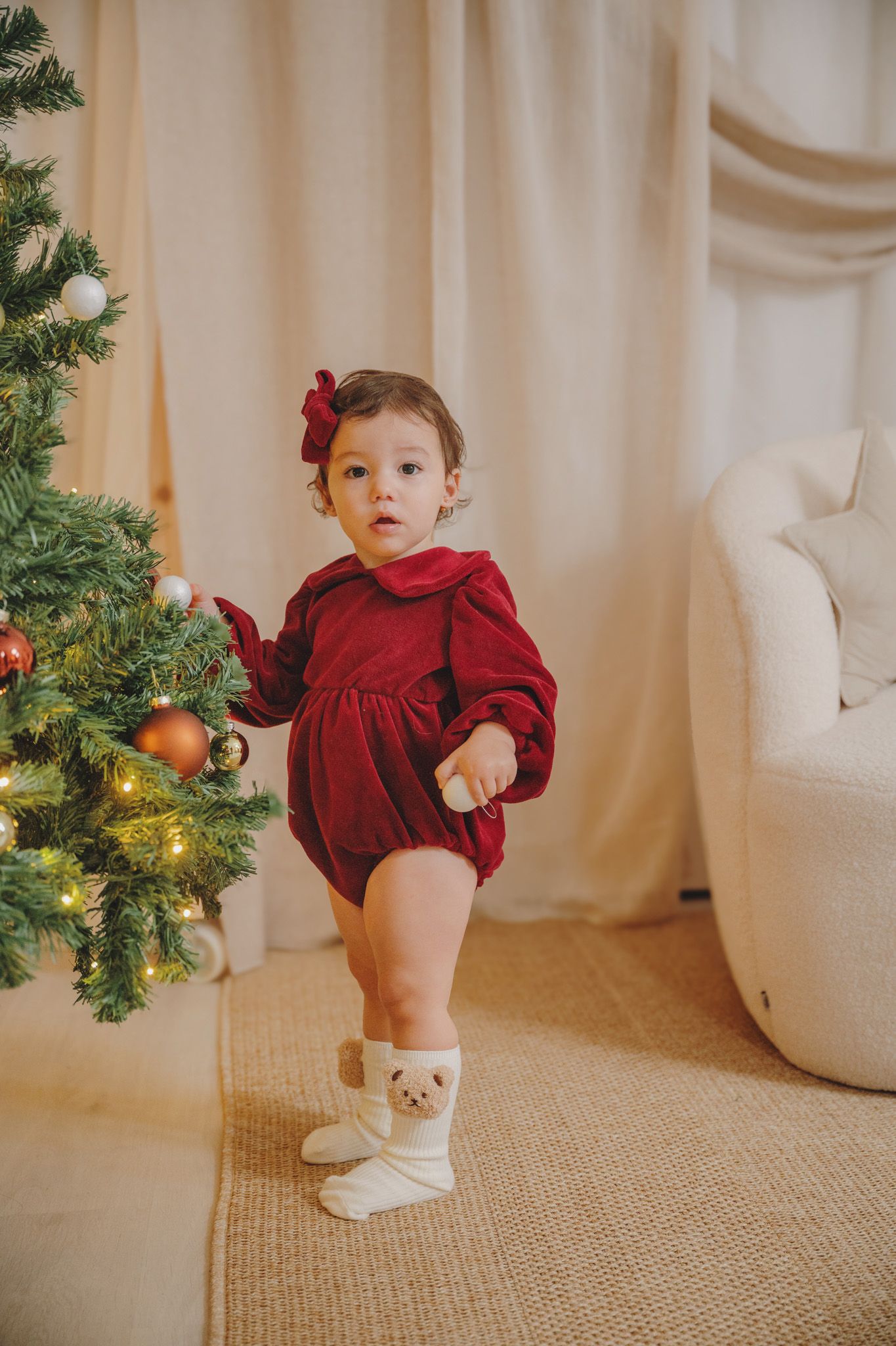 Toddler in a red velvet romper and matching hair bow stands in teddy bear socks beside a decorated Christmas tree in a cozy cream room.