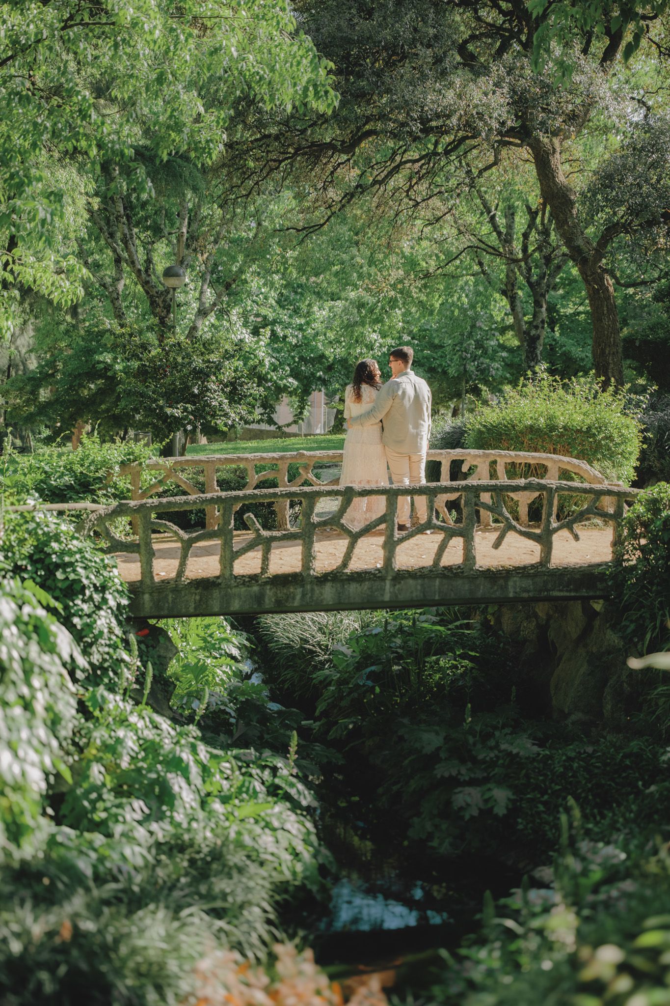Romantic couple walking across a wooden bridge surrounded by lush green trees.