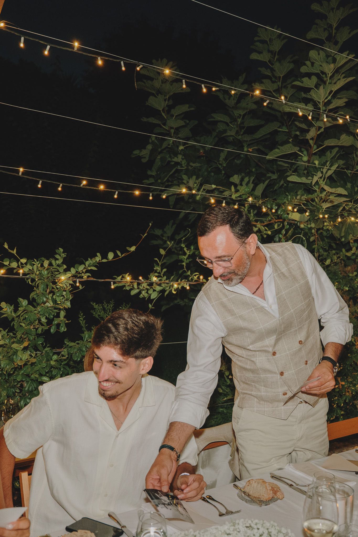 Guests at an outdoor evening reception sit at a long table under string lights.